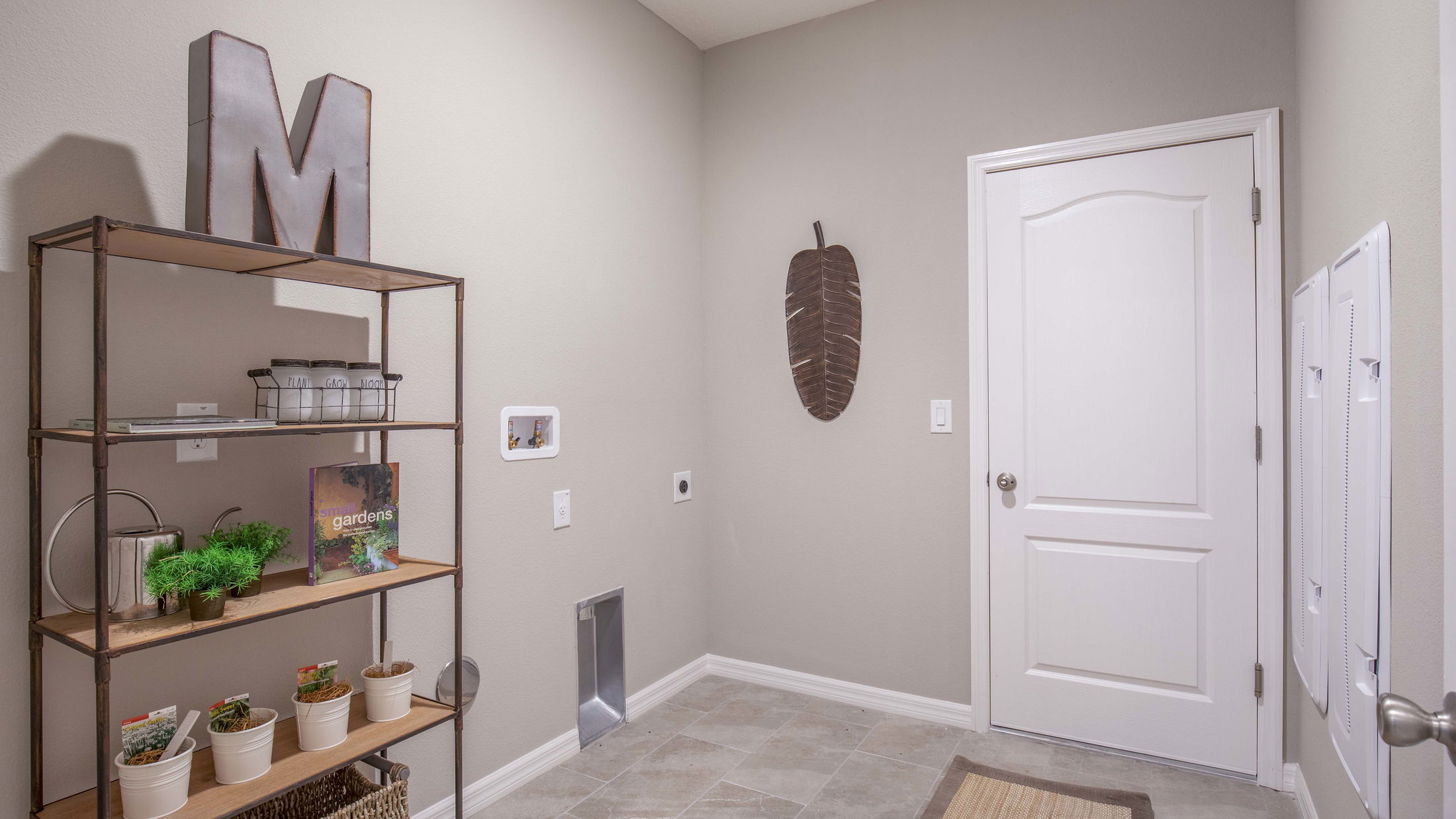 Modern laundry room with metal shelving, decorated with gardening books and plants, next to a white door and wall art.