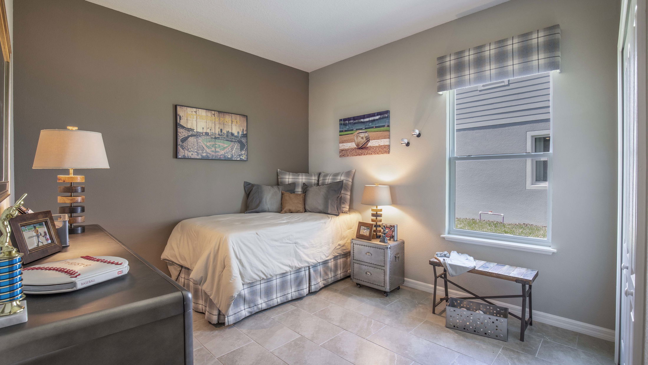 Cozy bedroom with baseball-themed decor, featuring a neatly made bed, wall art, and a sunlit window with an outdoor view.