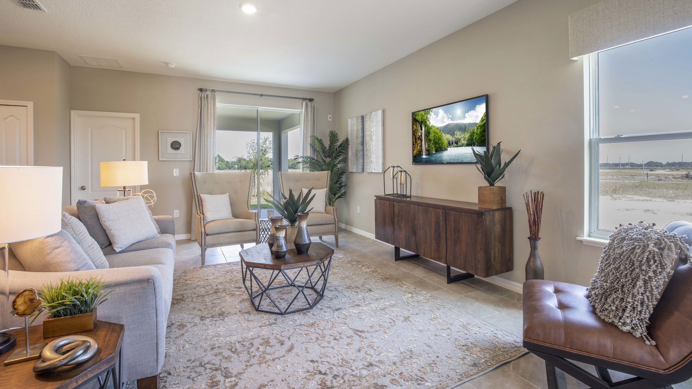 Elegant modern living room with neutral tones, featuring a stylish sofa, armchairs, decorative plants, and a wall-mounted TV displaying a scenic view.