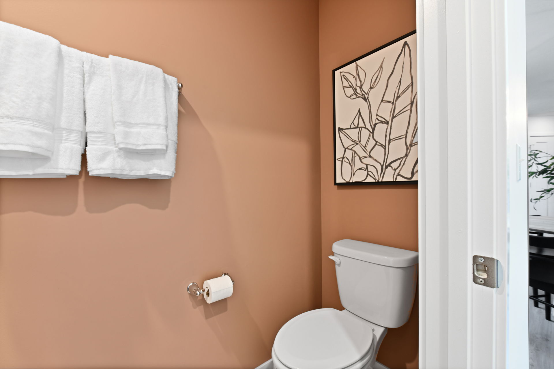 Contemporary bathroom with terracotta walls, featuring white towels, a toilet, and botanical artwork.