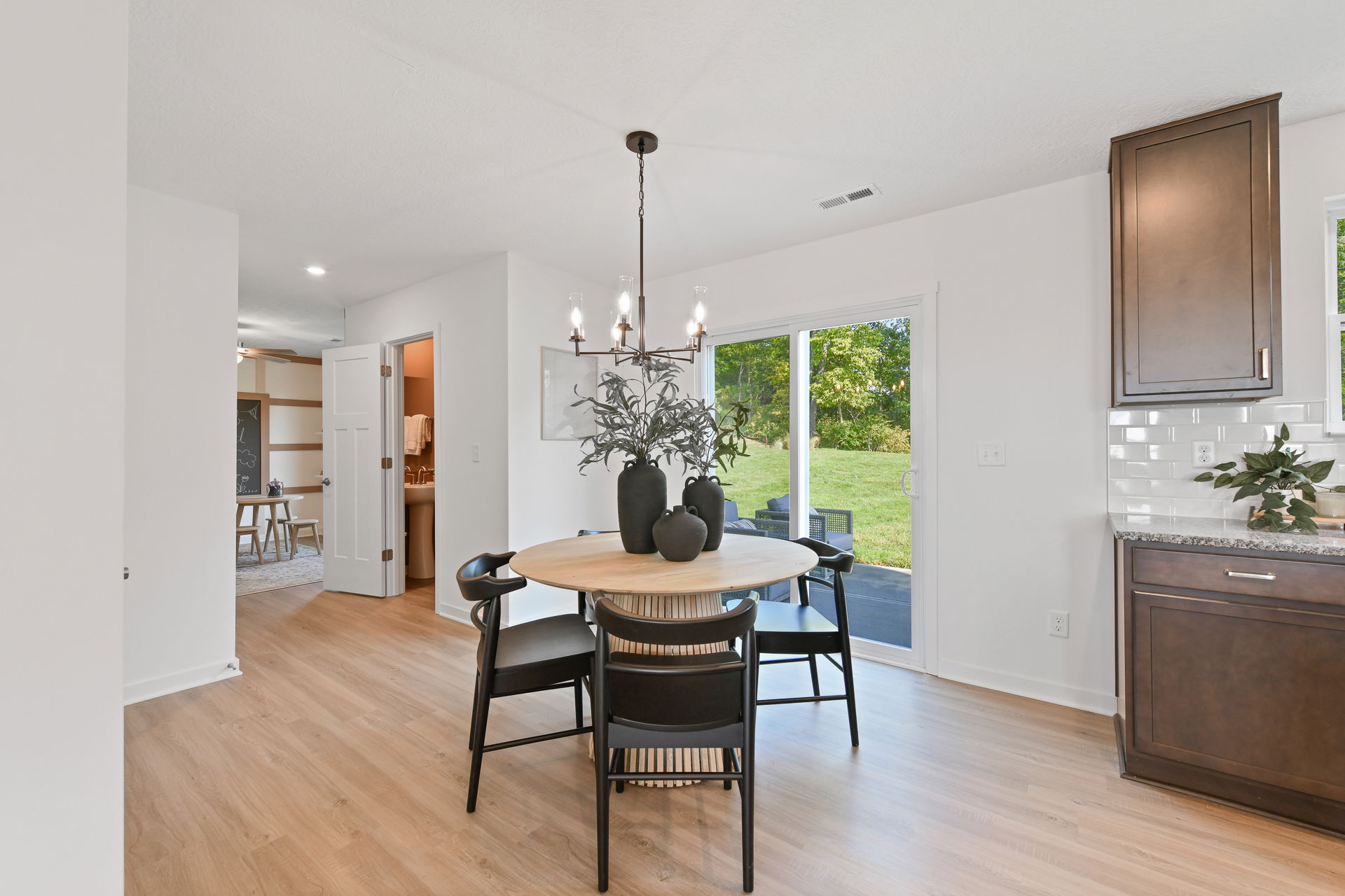 Modern dining room with a round wooden table, black chairs, elegant chandelier, and a view of a lush backyard through sliding glass doors.