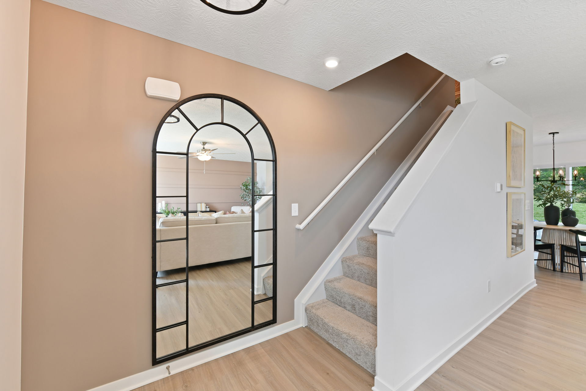 Stylish modern entryway with a decorative arched mirror, beige walls, and a staircase beside a dining area.