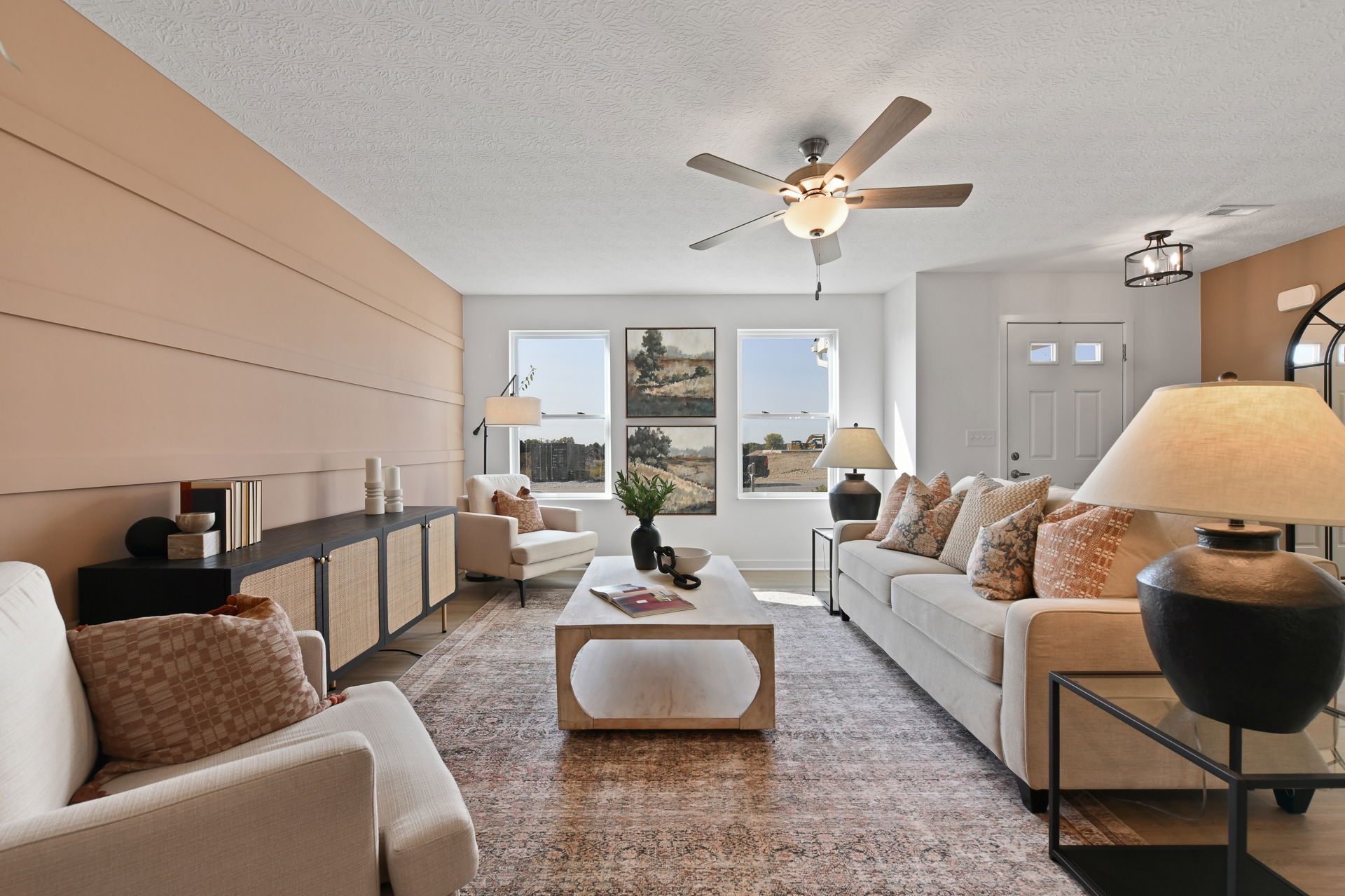 Modern living room with beige sofas, patterned rug, ceiling fan, and large windows showcasing outdoor views.