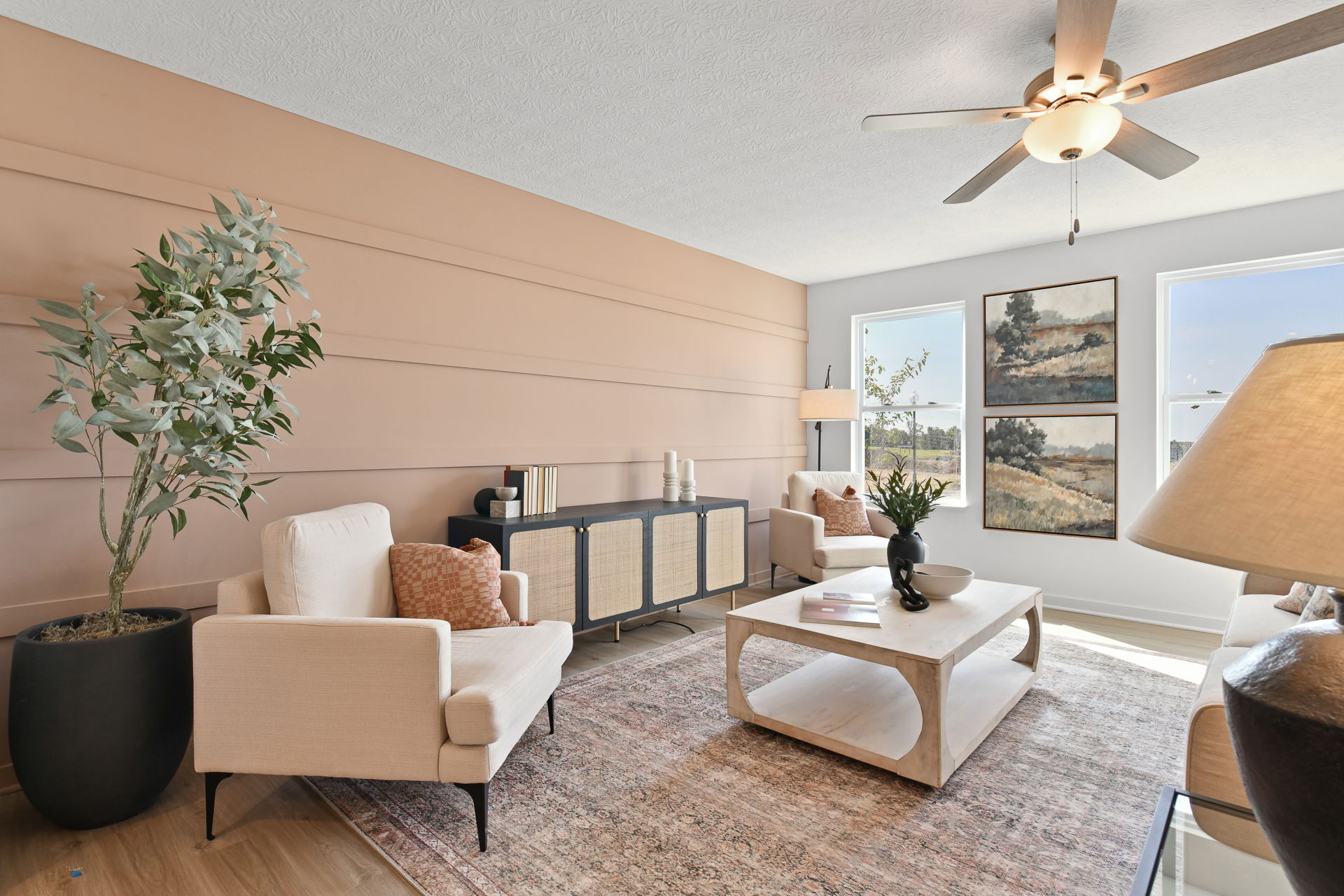 Modern living room with beige furniture, a light wood coffee table, decorative artwork, and a ceiling fan, bathed in natural light.