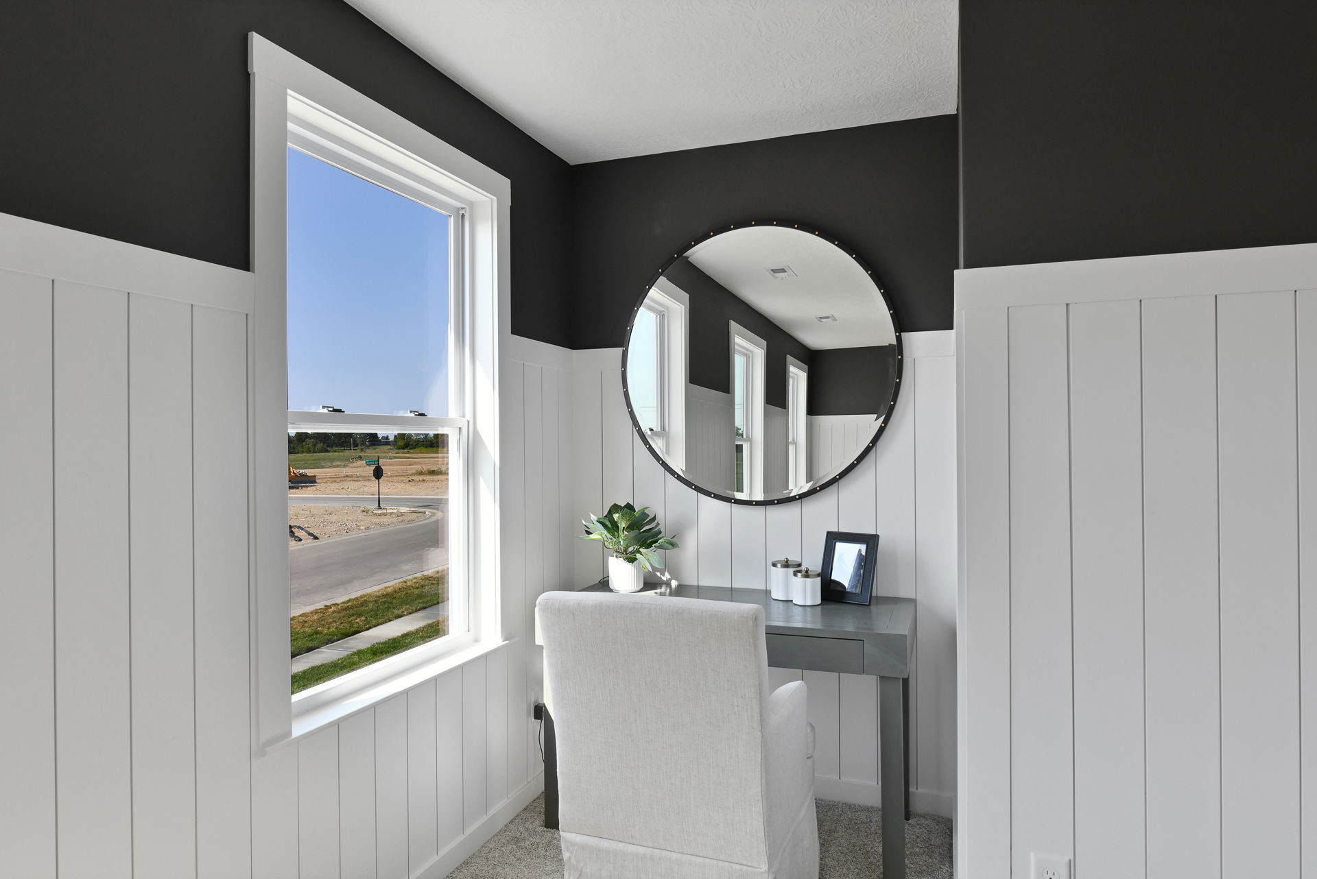 Stylish home office corner with a modern desk, cushioned chair, large round mirror, and a scenic window view.