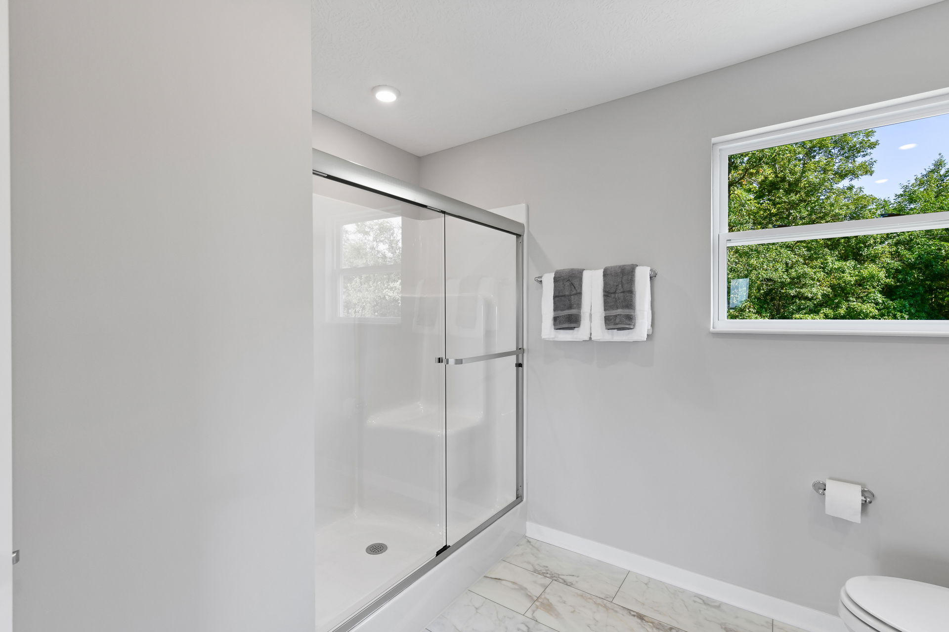 Modern bathroom with a glass-enclosed shower, double towel rack, and large window overlooking vibrant greenery.