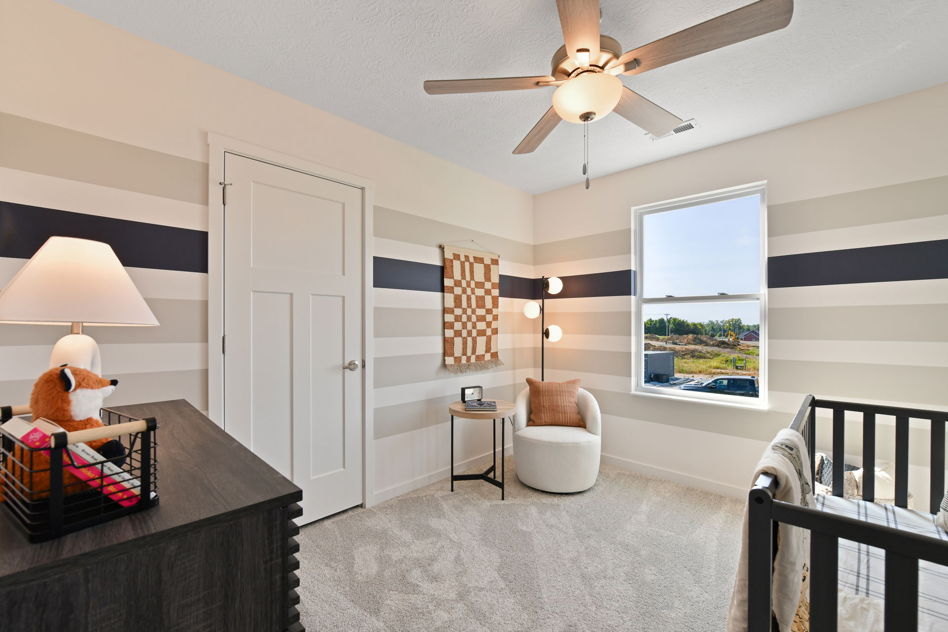 Modern nursery room with striped walls, a ceiling fan, cozy chair, and plush decor in a bright, inviting space.