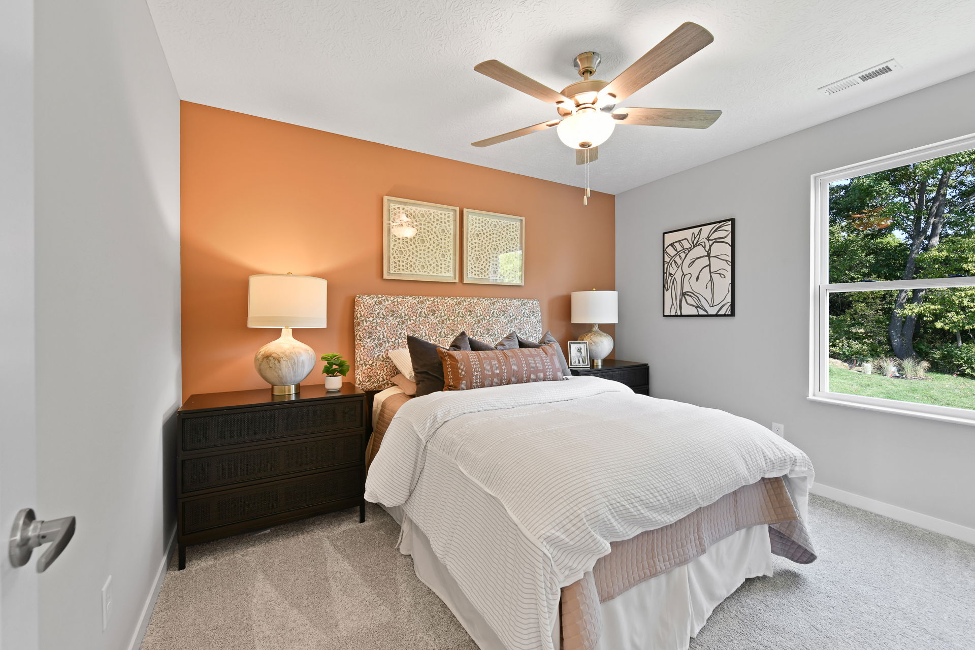 Modern bedroom with orange accent wall, cozy bedding, elegant décor, and a ceiling fan, overlooking a lush outdoor view.