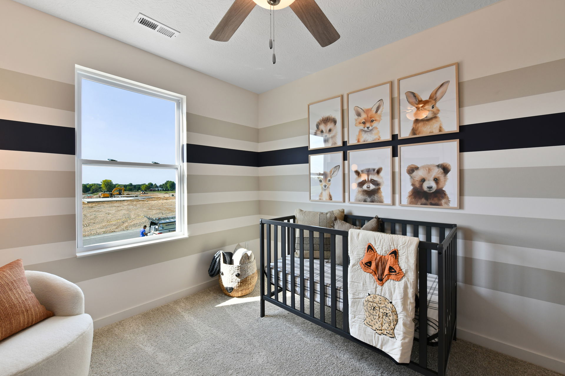 Modern nursery room with striped walls, animal artwork, a black crib, and a view of a construction site outside the window.