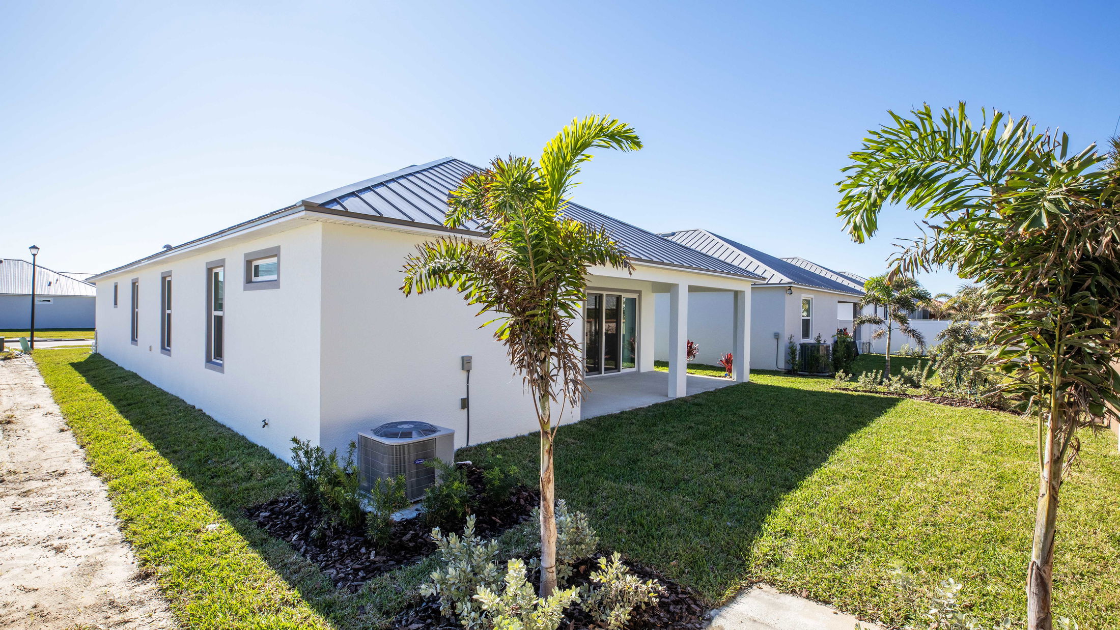 Modern white house with a metal roof and a landscaped backyard featuring palm trees under a clear blue sky.