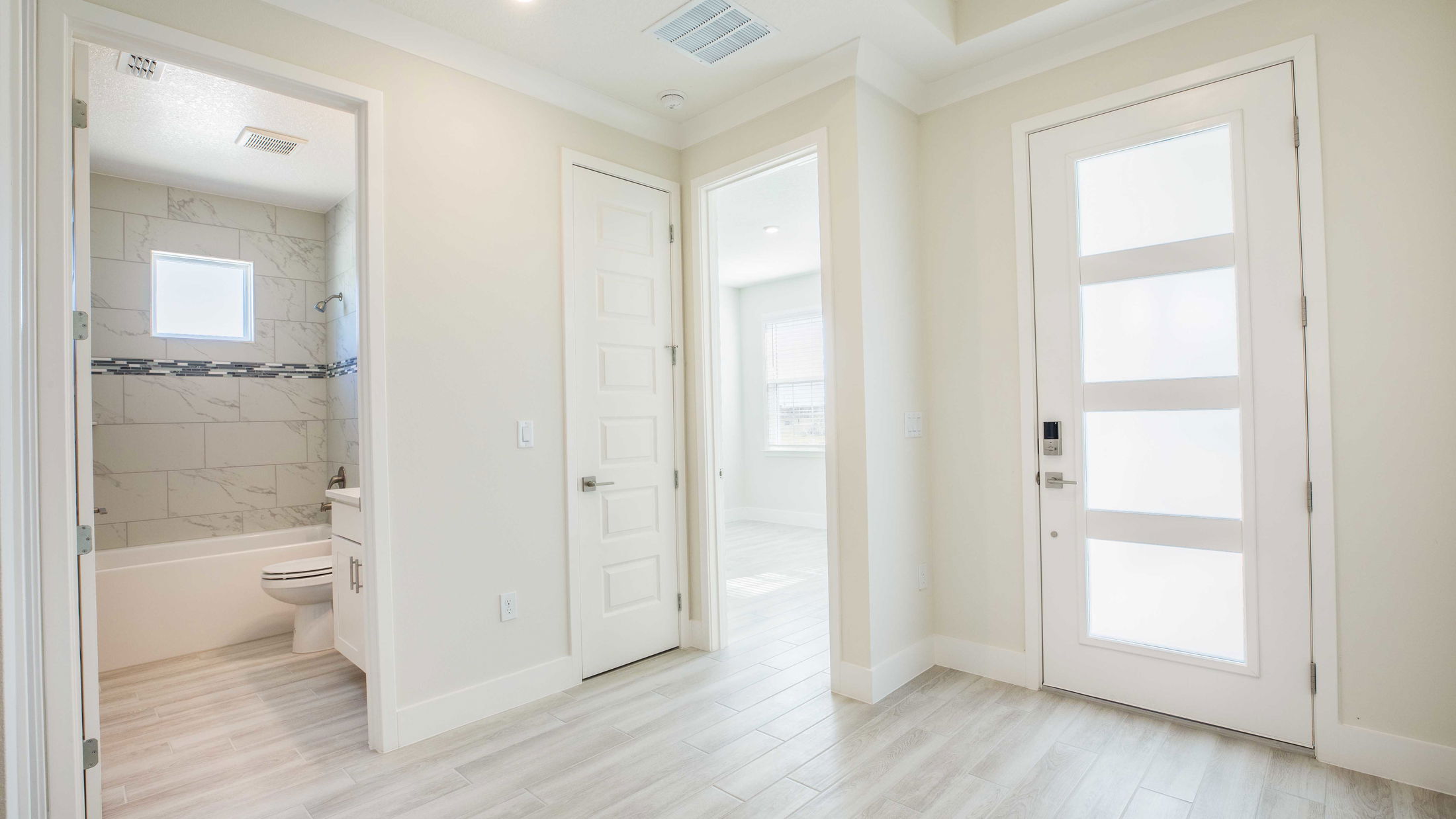 Modern interior showing a bright hallway with light wood flooring, a bathroom with marble tiles, and a frosted glass front door.