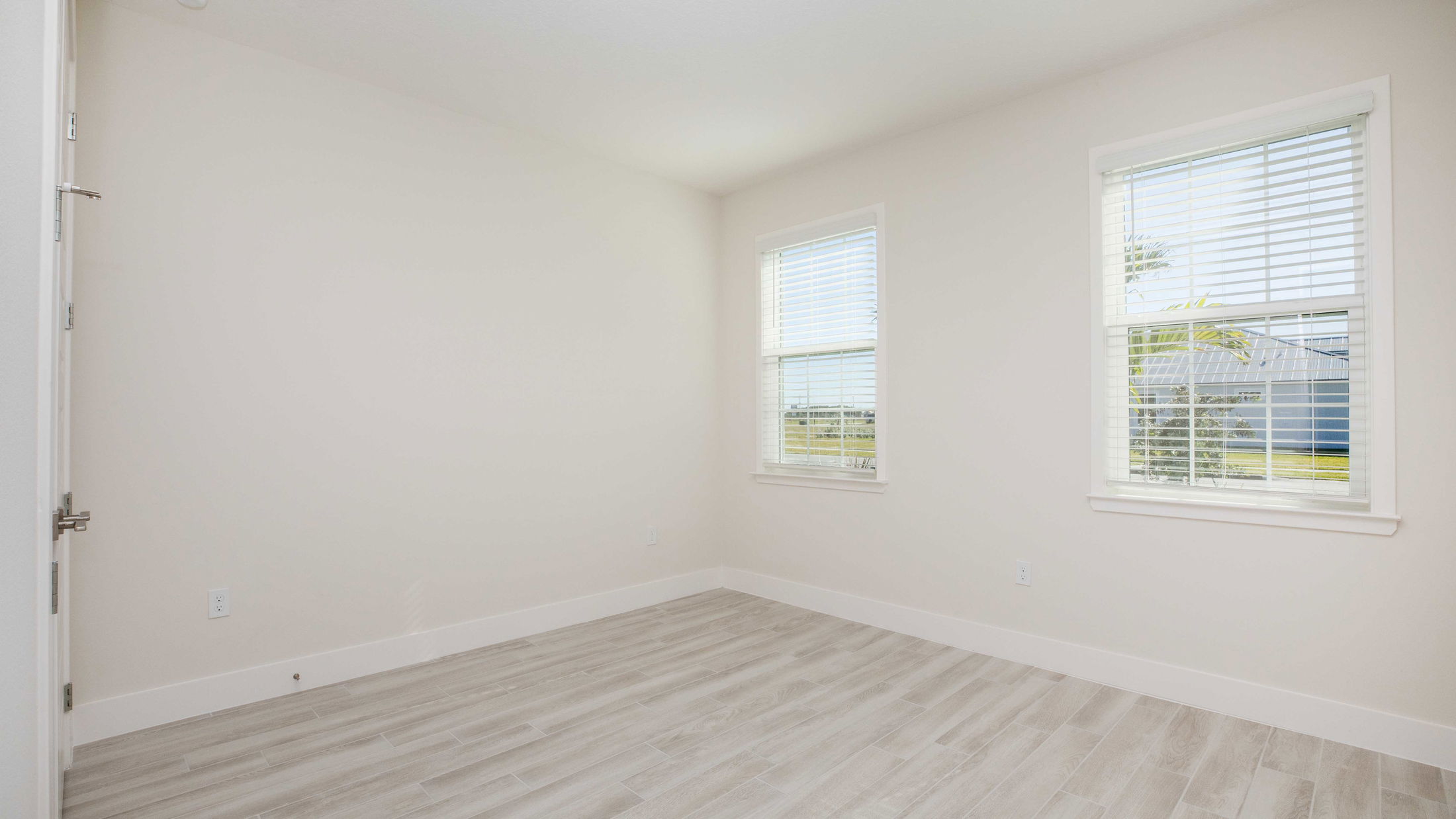 Bright, empty room with light wood flooring, white walls, and two windows with blinds.