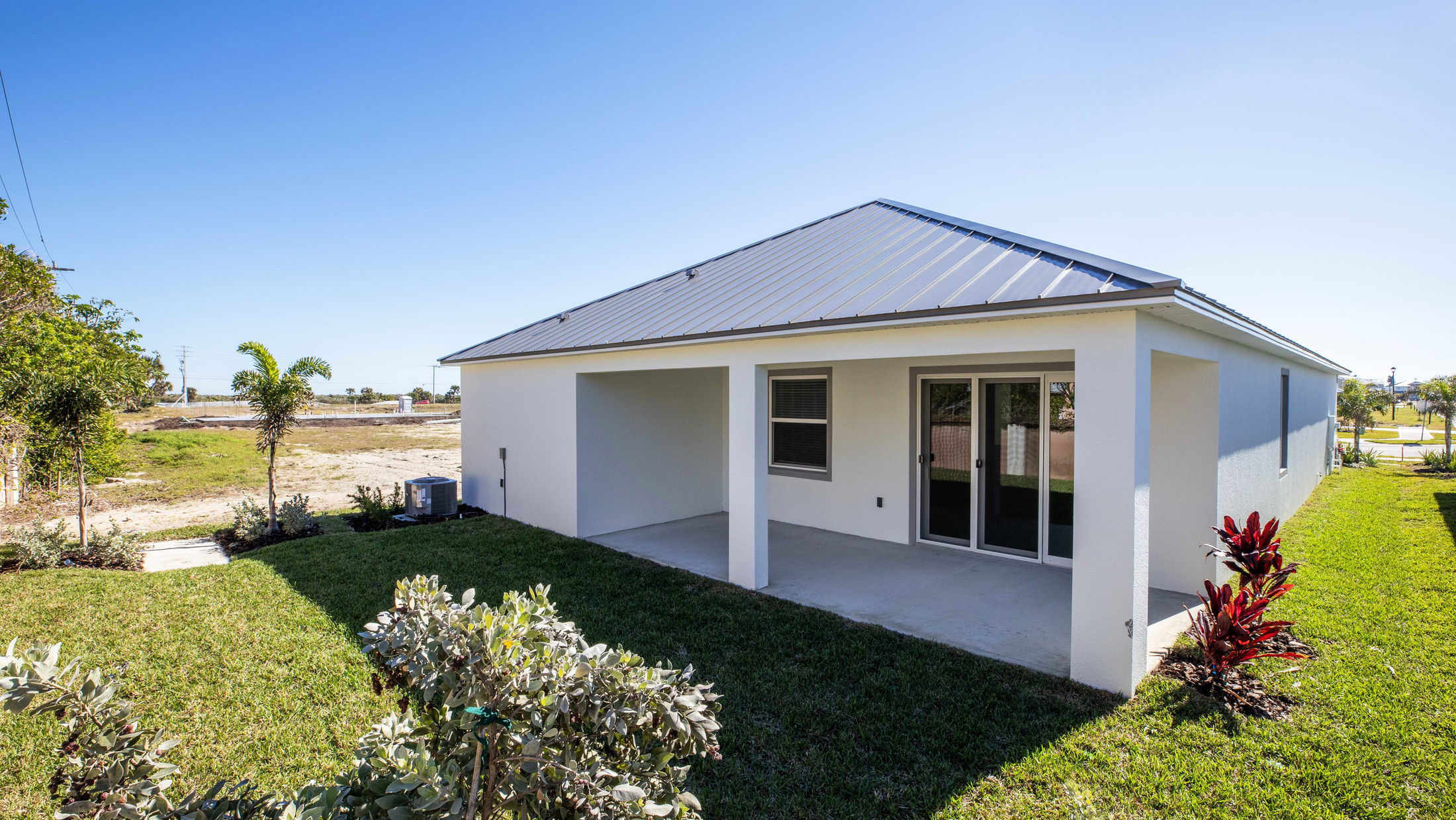 Backyard view of a modern white house with a metal roof and a small patio surrounded by a well-maintained lawn and tropical plants.