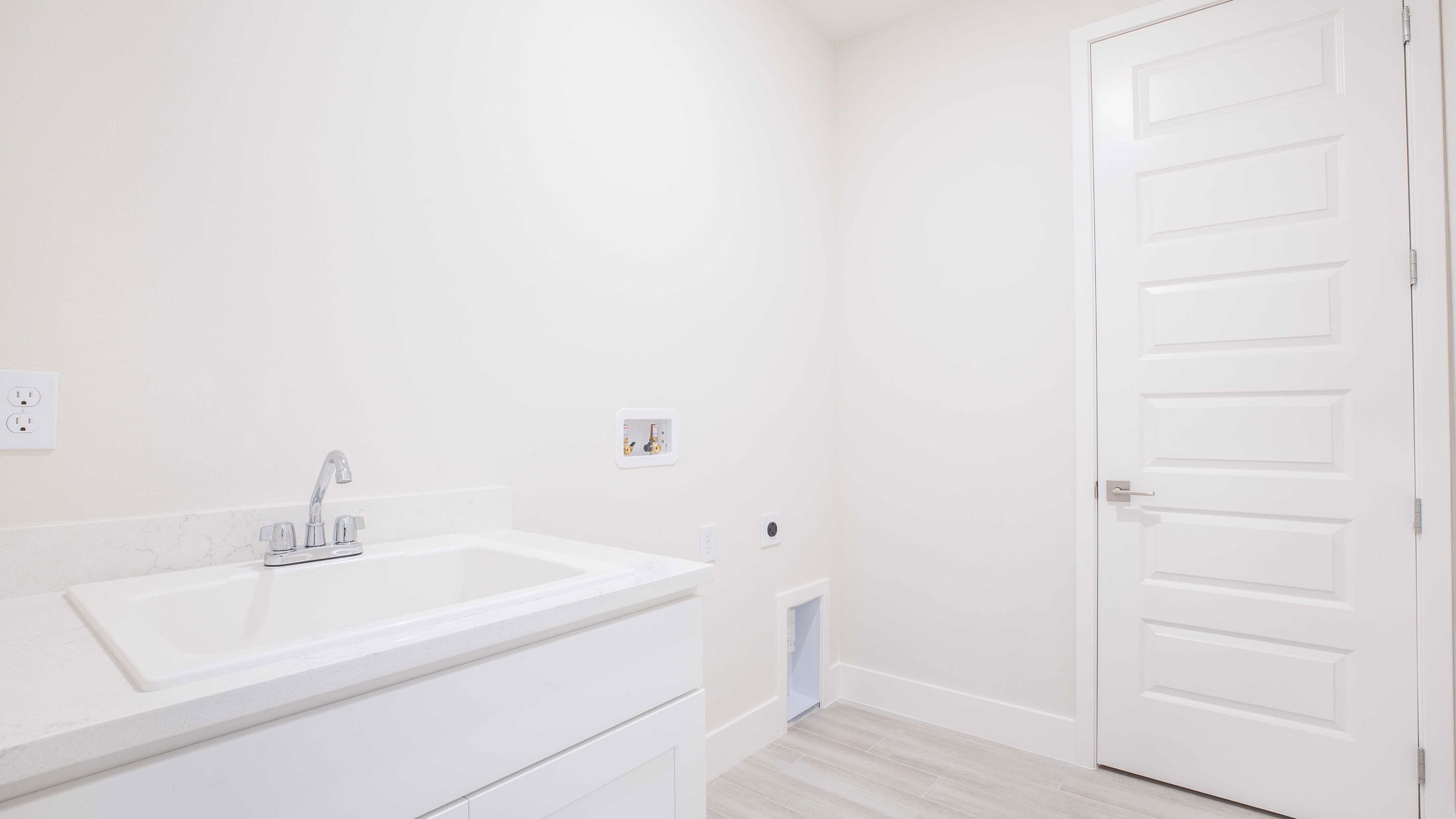 A bright and modern laundry room with a white utility sink, plumbing connections, and a door, featuring minimalist design.