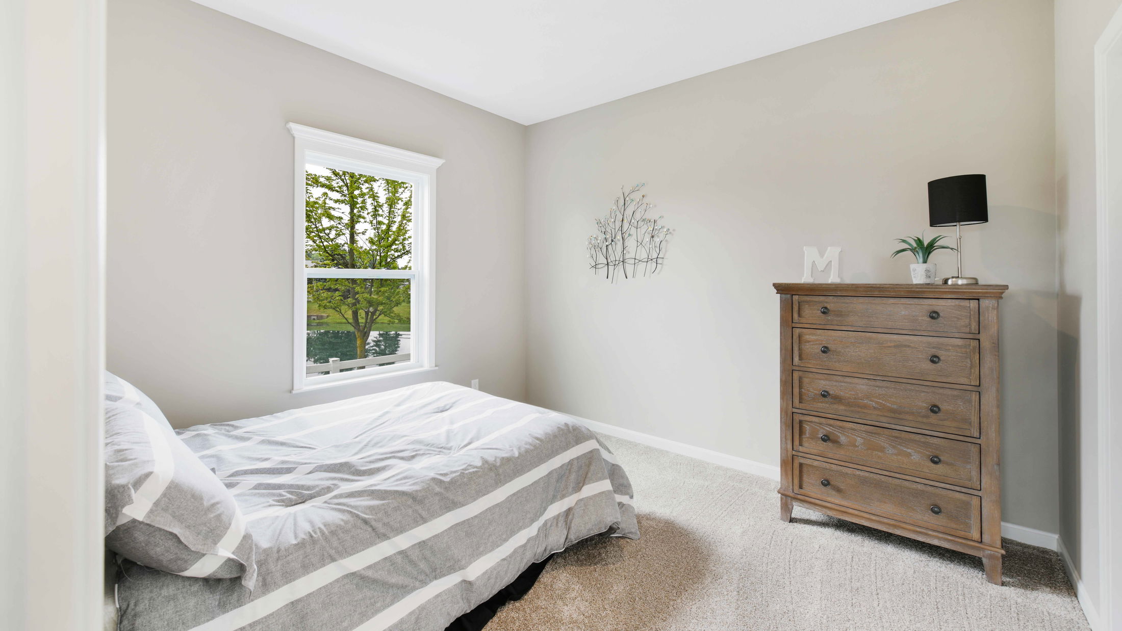 Minimalist bedroom with a gray and white striped bed, wooden dresser, and a window overlooking a green tree.