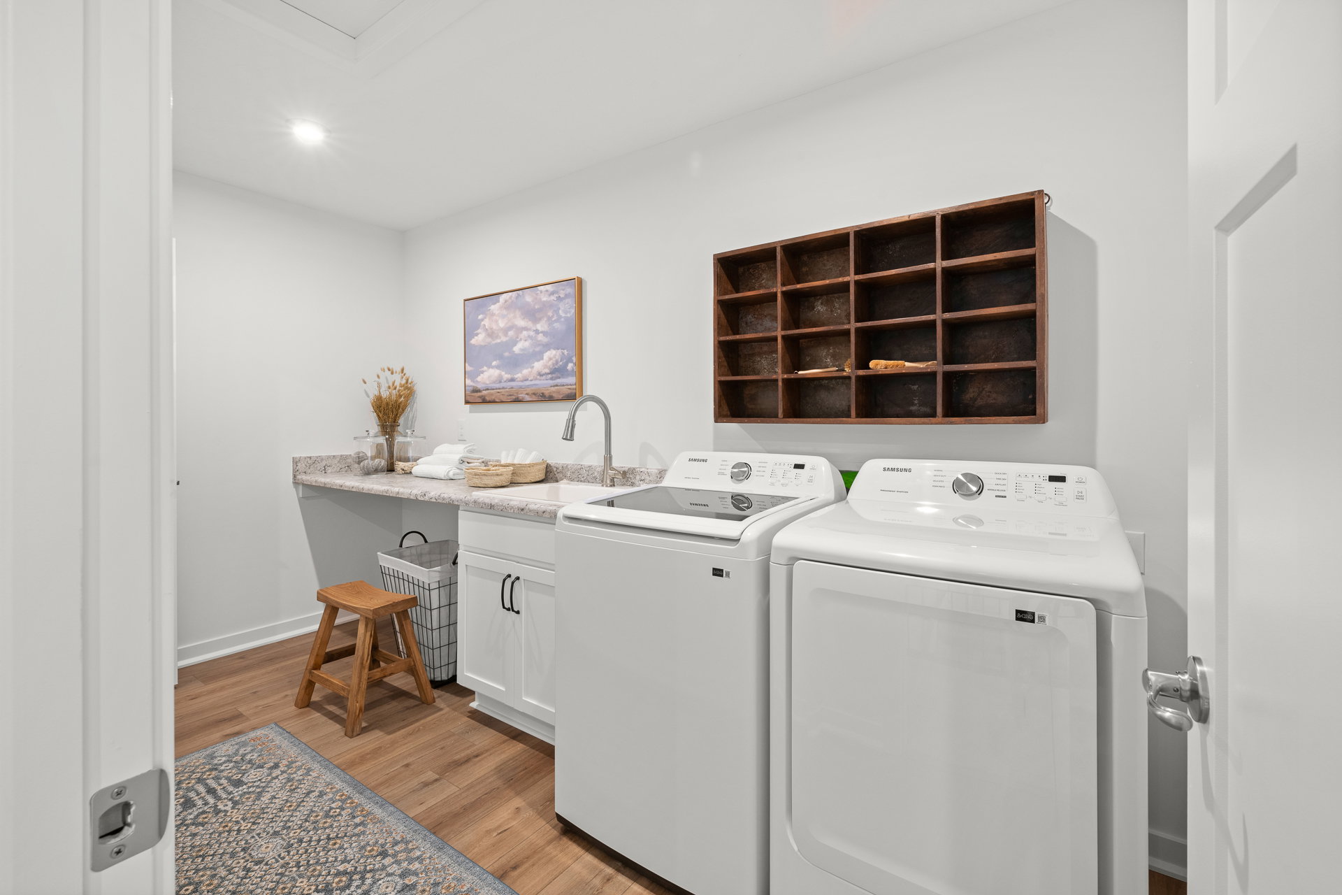A bright laundry room featuring two white washing machines, a countertop, a wooden shelf with decorative items, and a small stool.