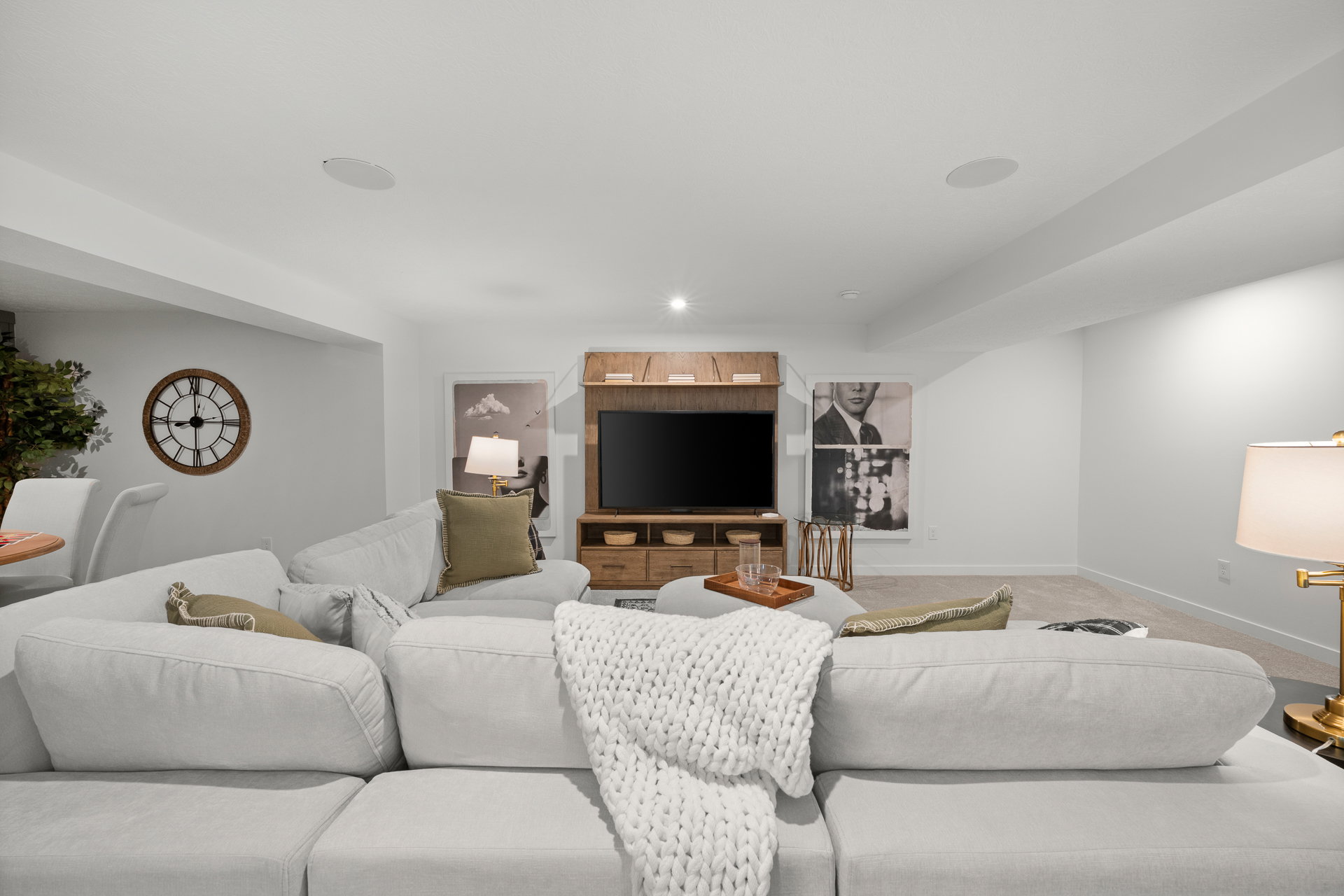 A cozy basement lounge area featuring a light-colored sectional couch, a wooden entertainment unit with a TV, and decorative wall art.