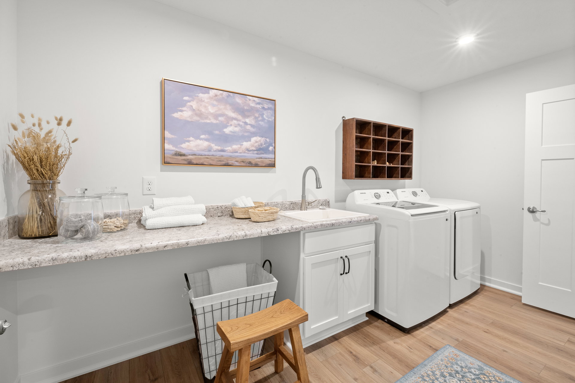 A bright laundry room featuring white appliances, a granite countertop, a wooden stool, and decorative storage with fresh flowers.