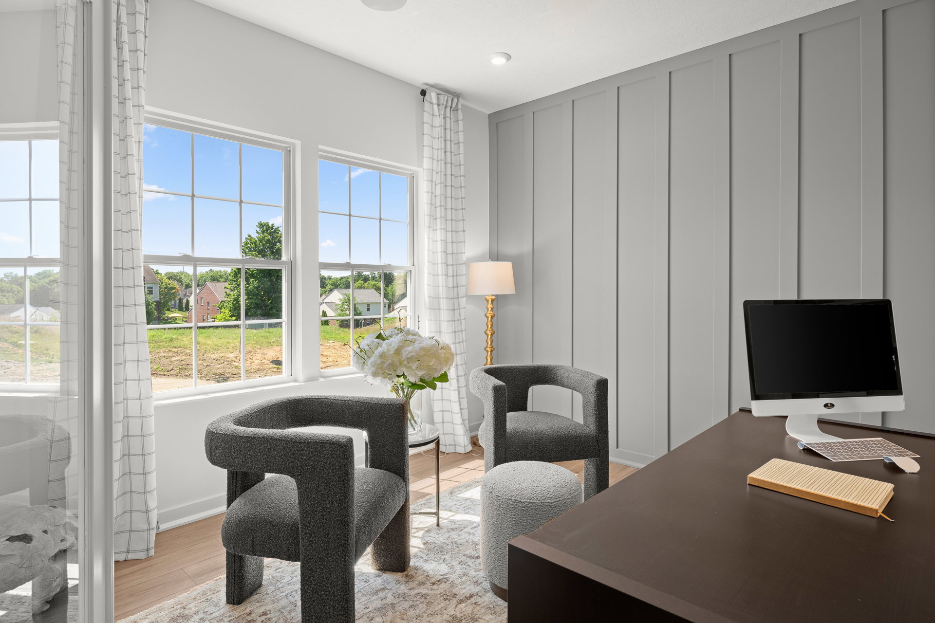 A modern office space featuring two upholstered chairs, a small round ottoman, a desk with a computer, and large windows with a view of a green landscape.