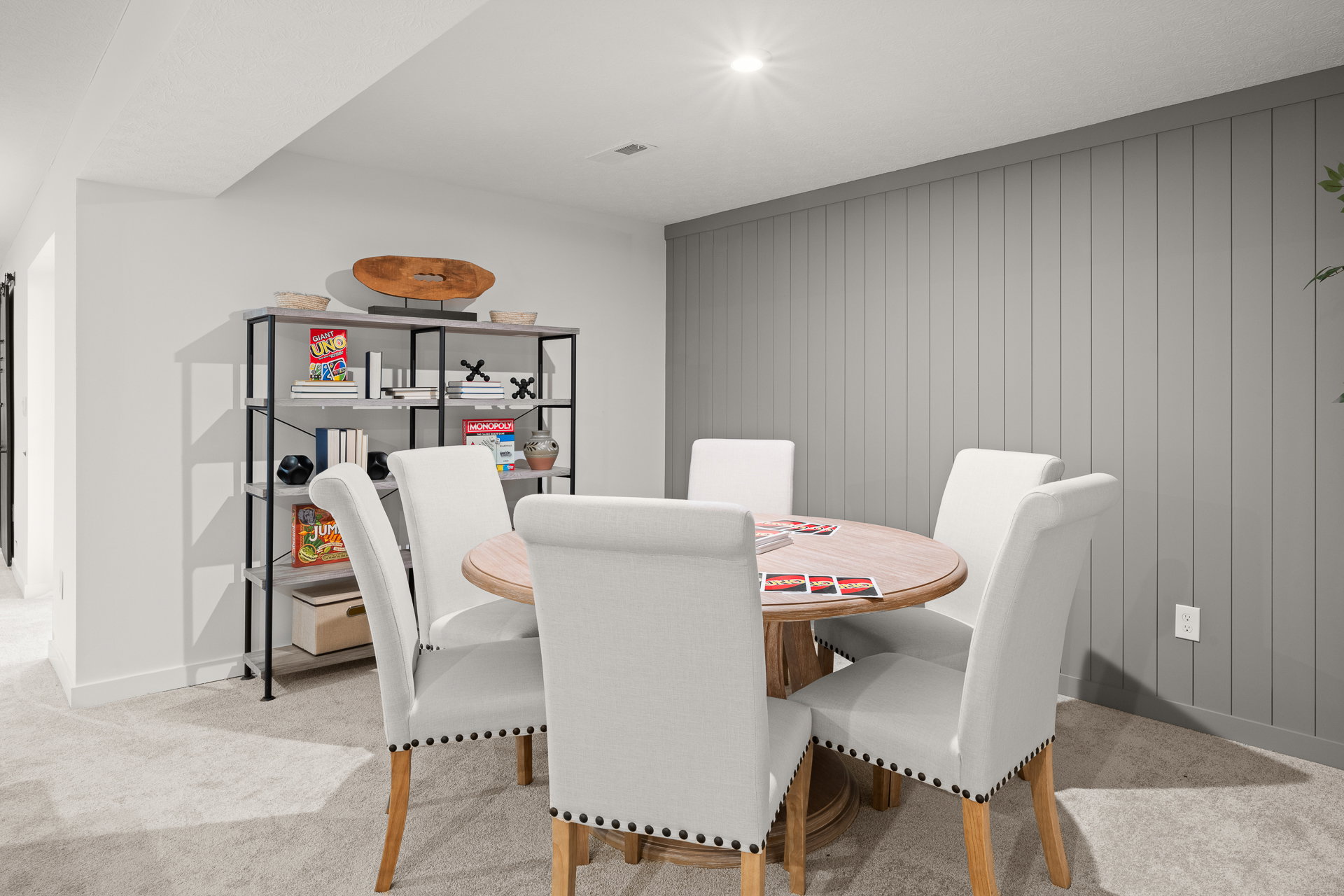 A round wooden dining table surrounded by five upholstered chairs in a modern interior space with gray walls and a black shelf displaying various items.
