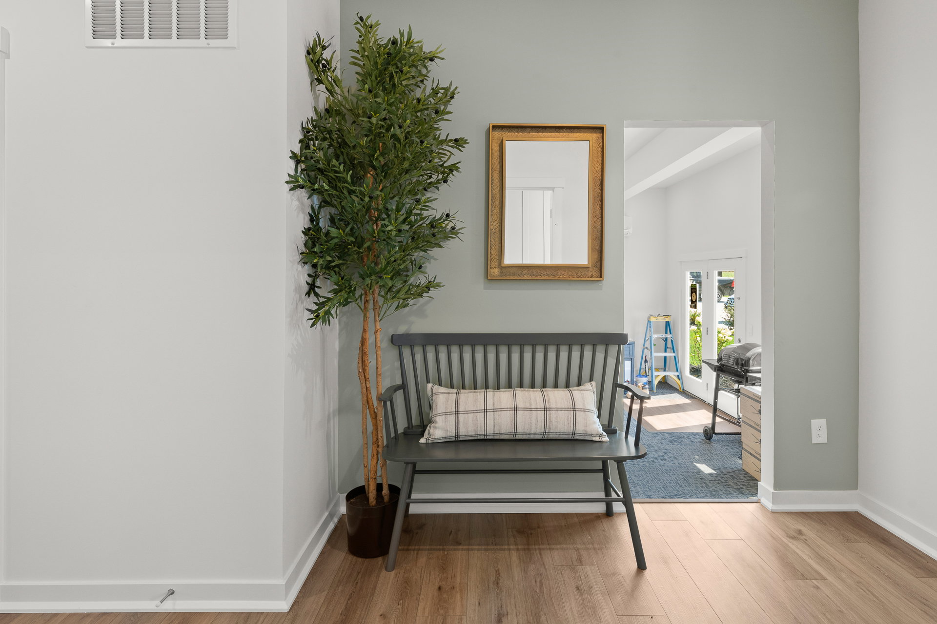 A gray bench with a cushion is positioned against a light green wall, next to a potted plant and a gold-framed mirror.