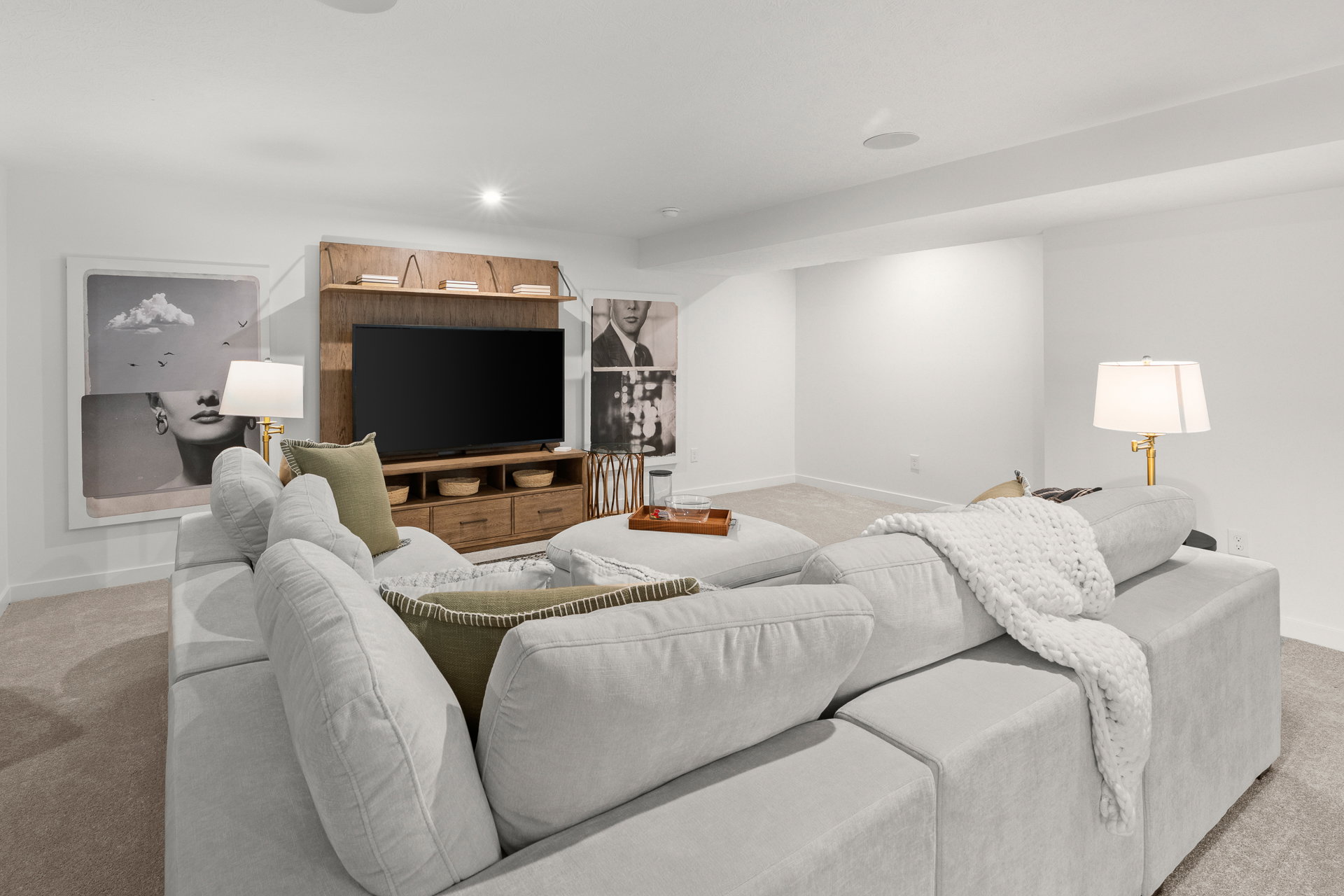 A cozy basement living area featuring a light gray sectional sofa, a wooden TV stand with a flat-screen TV, and decorative lamps, with artwork on the walls.