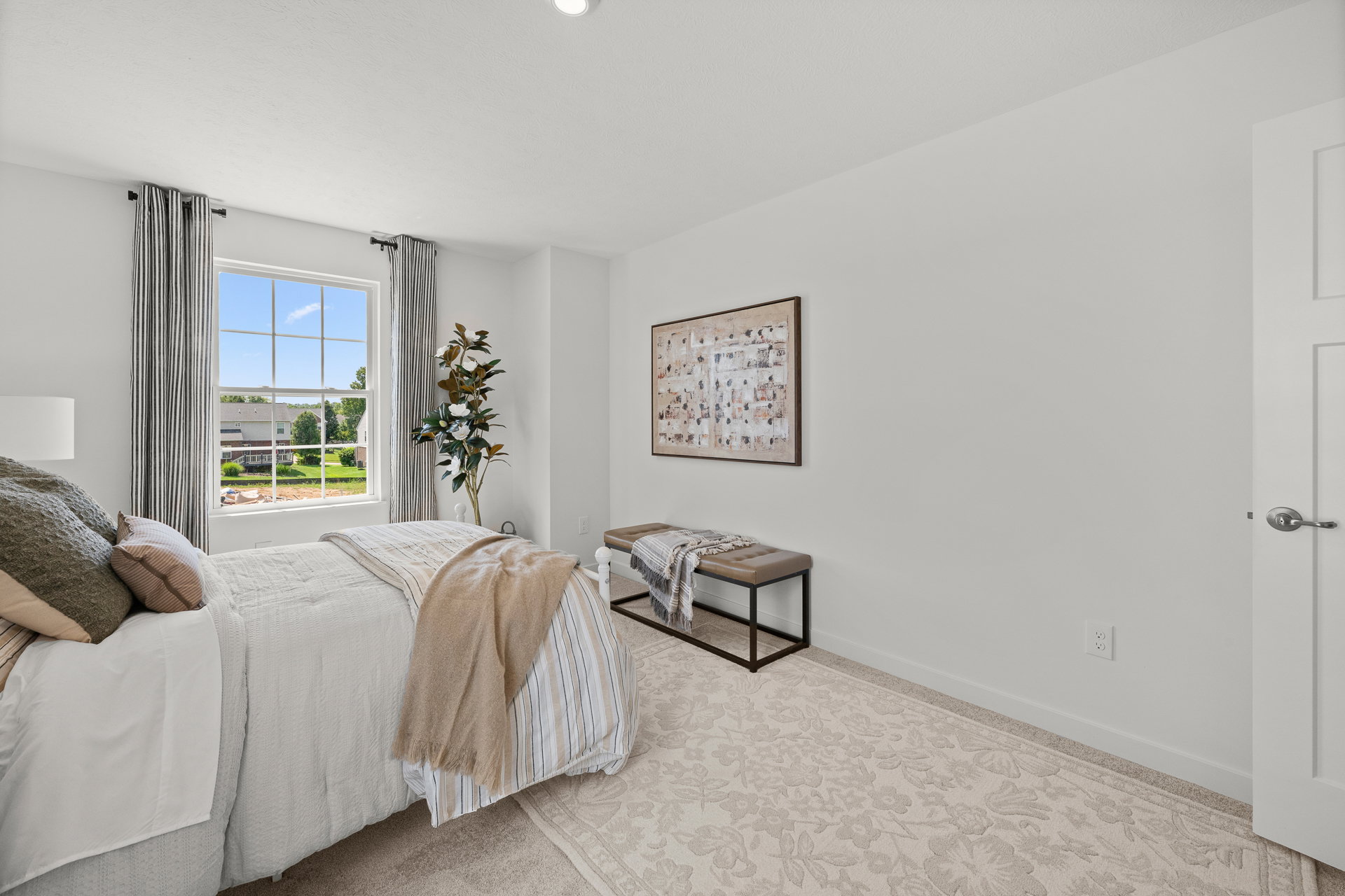 Bright bedroom featuring a bed with decorative pillows, a bench, and a window with a view of greenery outside.
