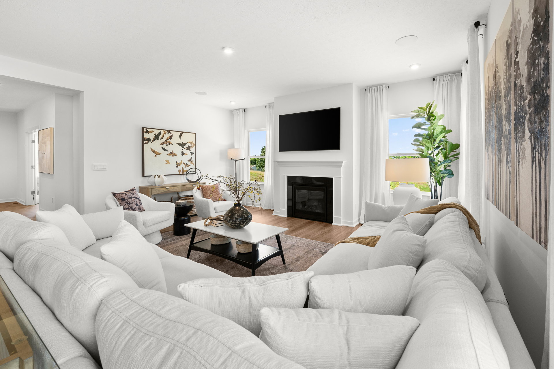 A modern living room featuring a white sectional sofa, a coffee table, a fireplace, and large windows with curtains, decorated with plants and artwork.