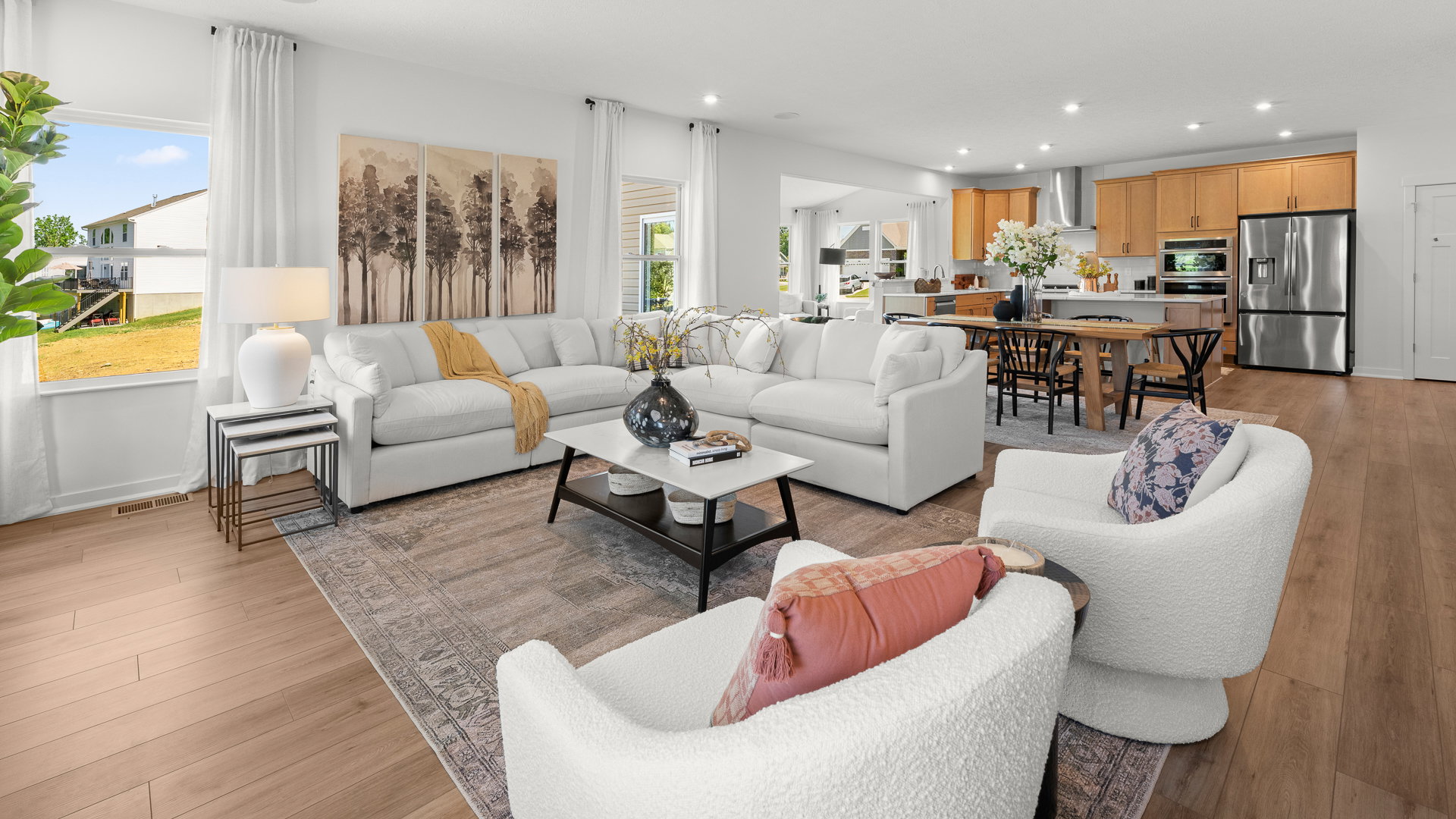 Bright, modern living room with a white sectional sofa, a coffee table, and large windows overlooking a green yard, featuring a nearby dining area and kitchen.