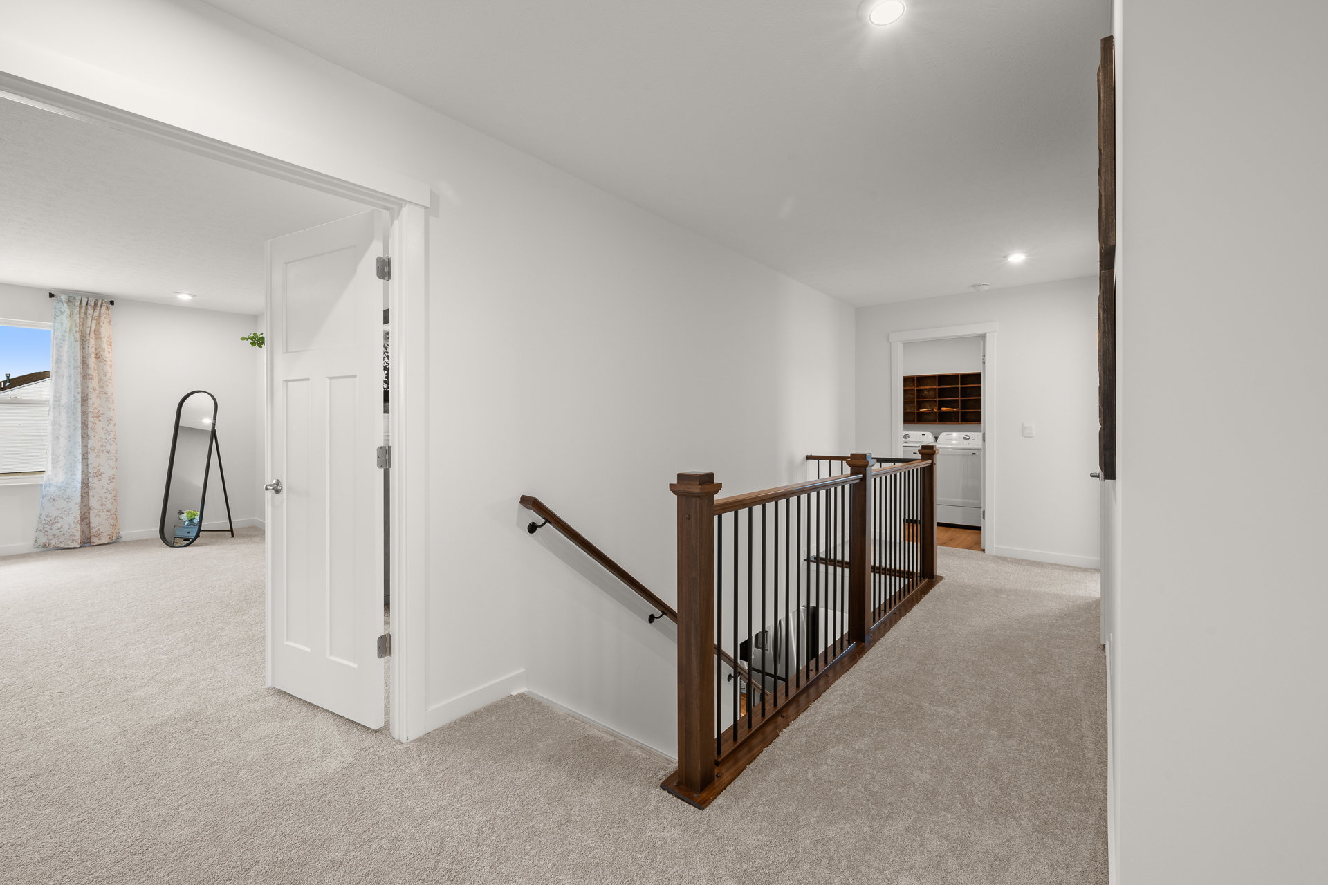 A well-lit hallway with carpeted floors, featuring a wooden railing and a door leading to a room.