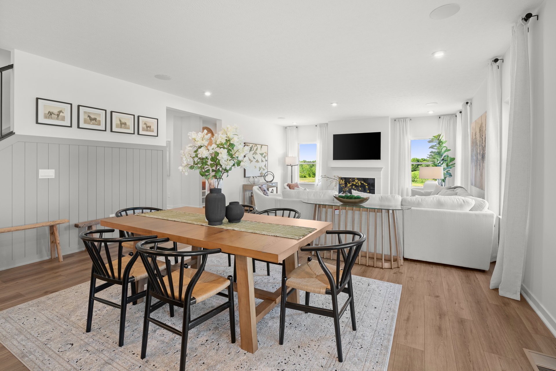 A modern dining area with a wooden table, black chairs, floral centerpiece, and a view of a living room featuring a sofa and a television.
