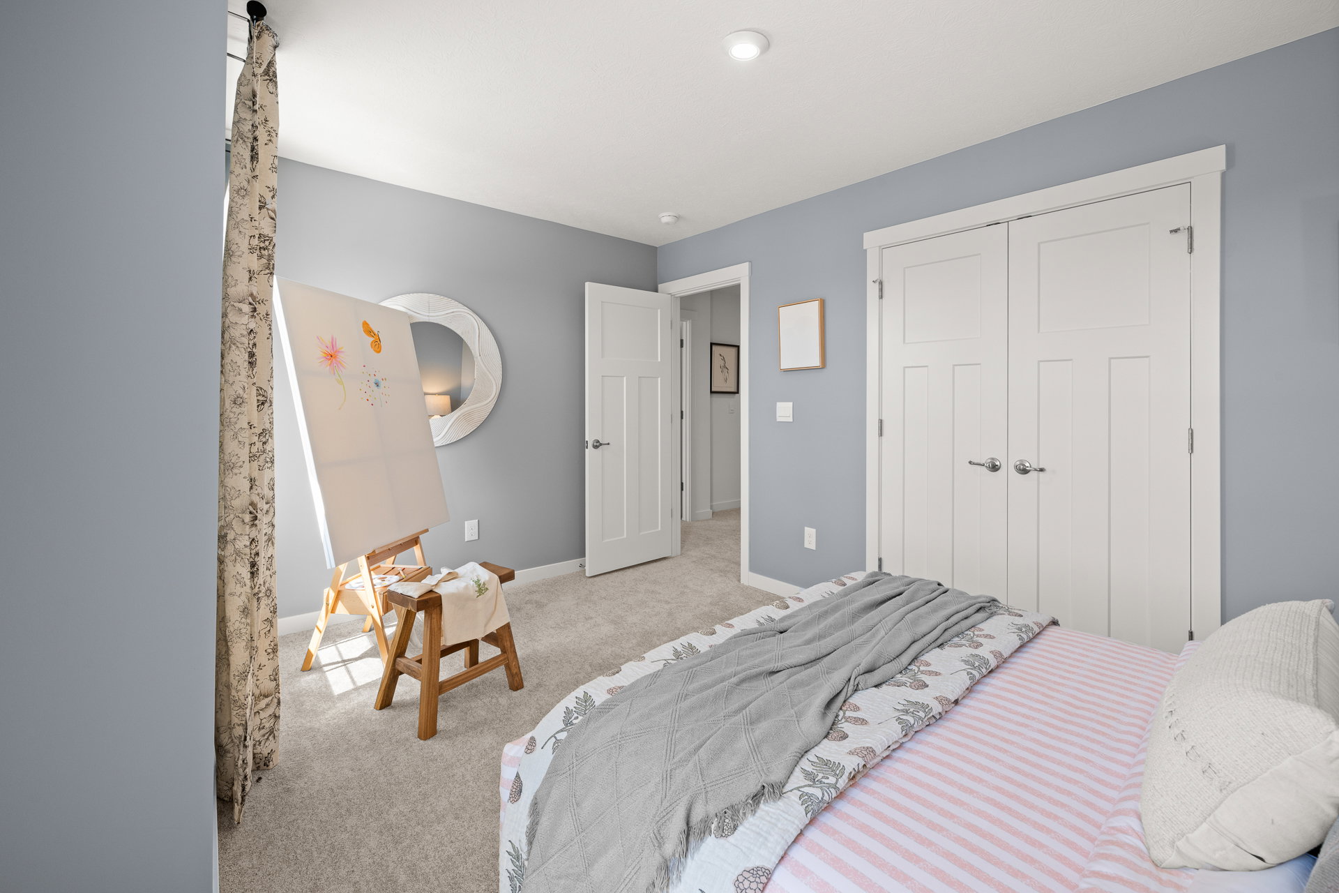 A well-lit bedroom featuring a bed with a patterned blanket, a small easel with artwork, and light gray walls.