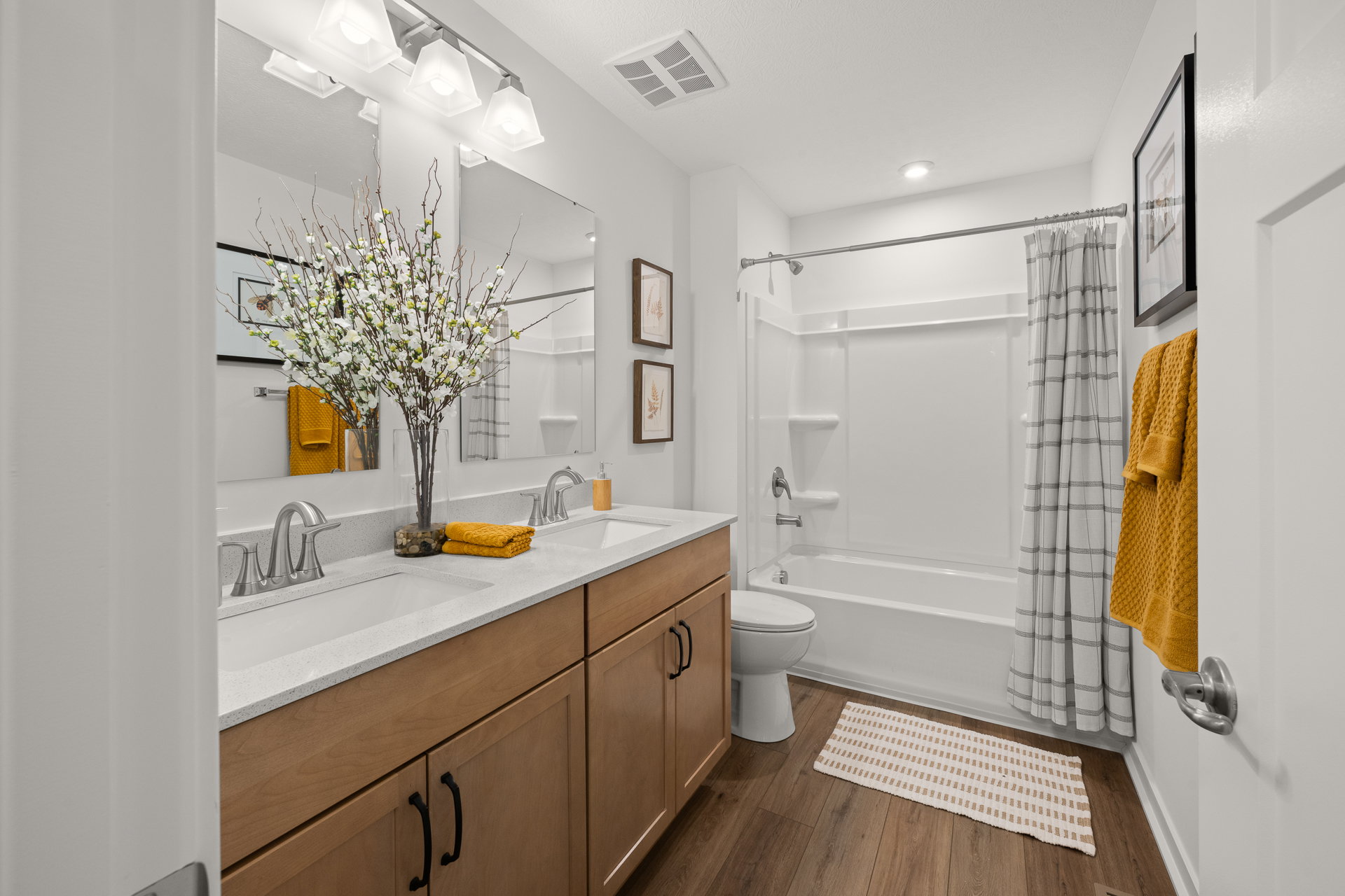 Bright bathroom featuring a double vanity with a quartz countertop, a bathtub with a shower curtain, and decorative elements like a floral arrangement and towels.