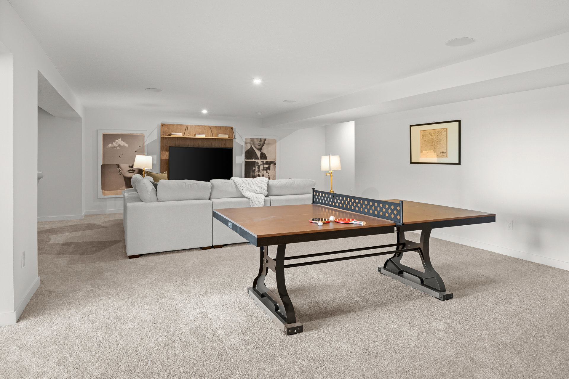 A spacious, well-lit basement recreation room featuring a ping pong table, a cozy sofa, and a flat-screen TV.
