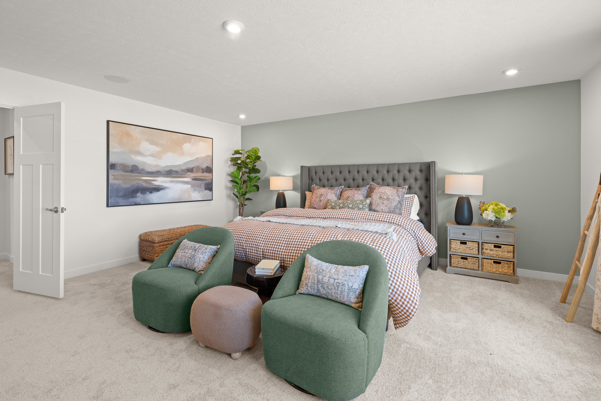 A cozy bedroom featuring a tufted gray bed, patterned bedding, green accent chairs, and a modern mural on the wall.