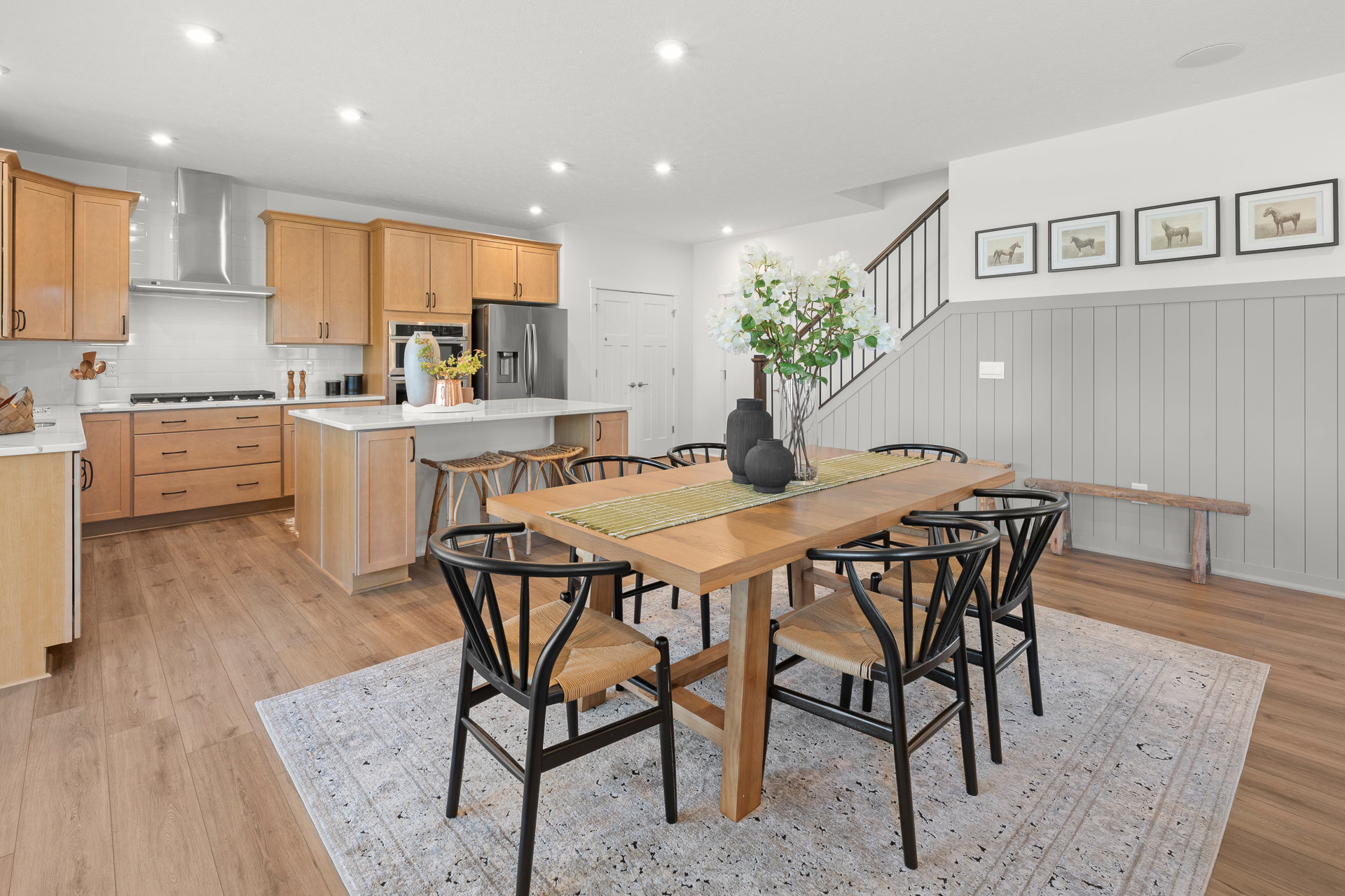 Modern dining area featuring a wooden table with six black chairs, a light-colored area rug, and a kitchen with wooden cabinets and stainless steel appliances.