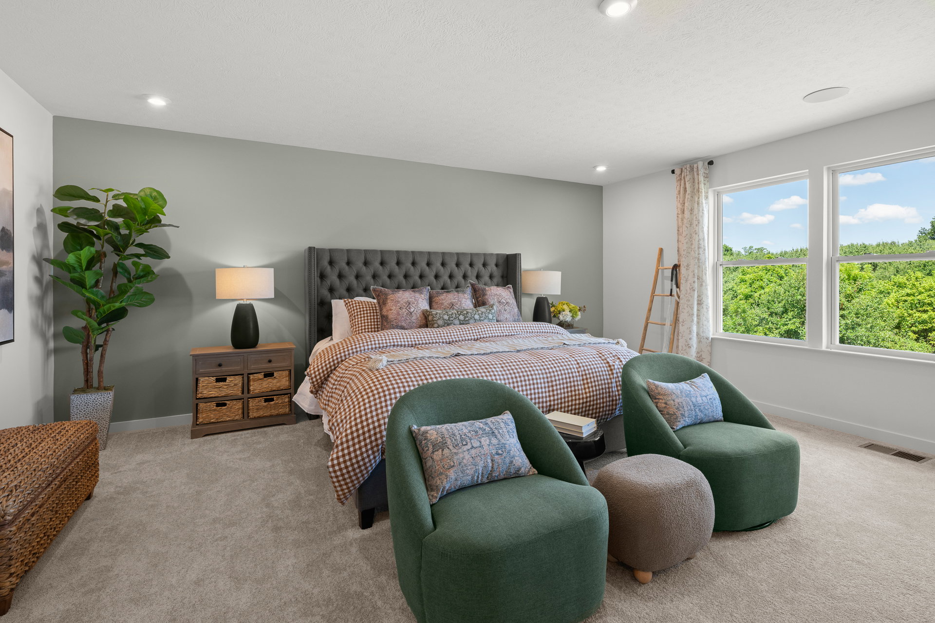 Modern bedroom featuring a large bed with patterned bedding, green accent chairs, a woven storage ottoman, and large windows overlooking greenery.