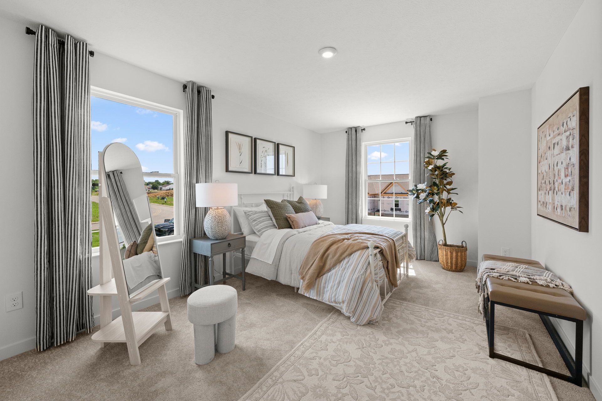 Bright, modern bedroom featuring a bed with striped bedding, a round mirror, a stool, and a view of the outdoors through large windows.
