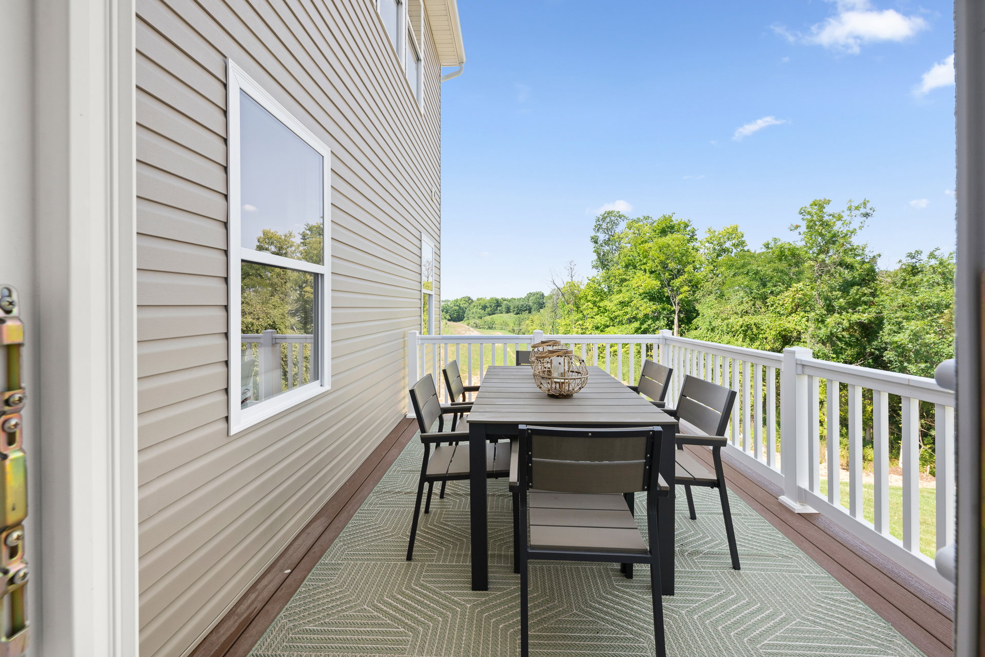 A large outdoor dining table with chairs on a balcony overlooking green trees and a clear blue sky.
