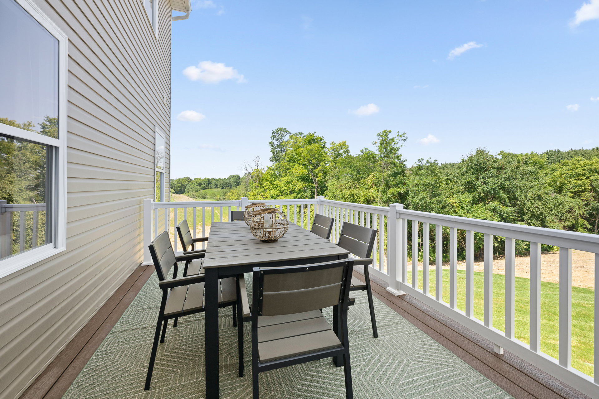 A spacious deck featuring a rectangular table and six chairs, overlooking green trees and a grassy area.