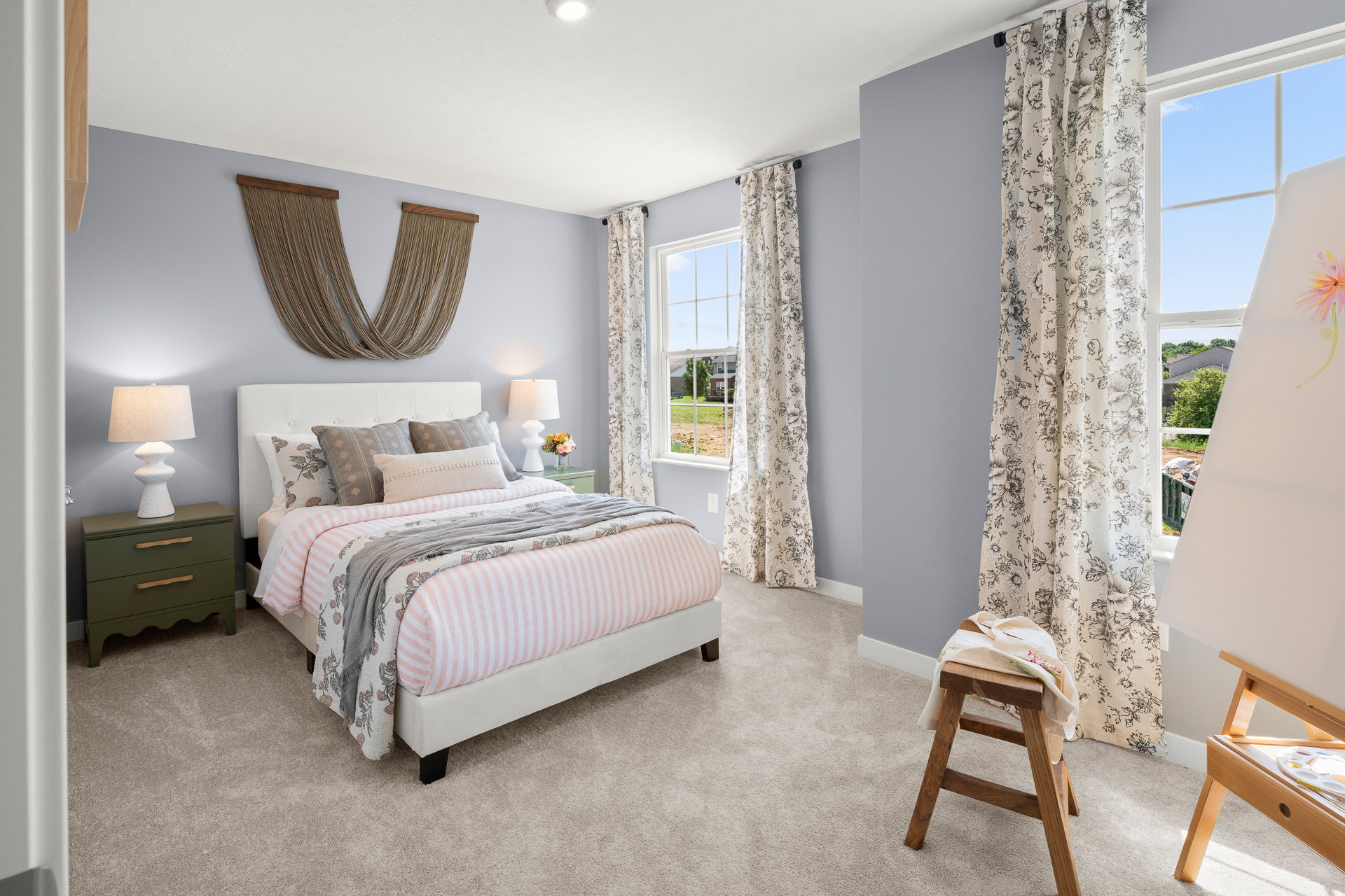 A cozy bedroom featuring a white bed with striped bedding, two large windows with patterned curtains, and a decorative wall piece above the headboard.