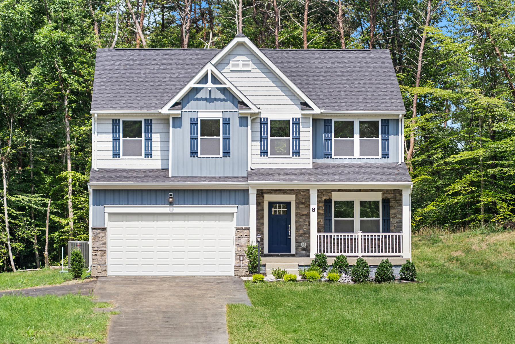 Modern two-story house with blue and white siding, stone accents, and a front porch, surrounded by lush greenery.