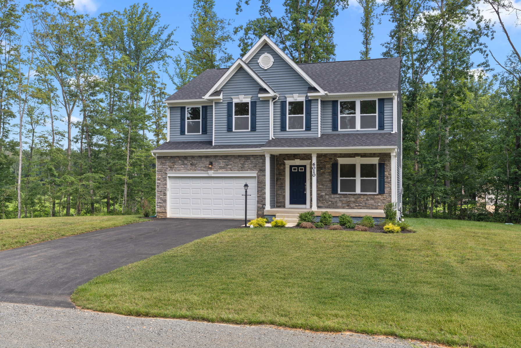 Modern two-story blue house with a stone facade and manicured lawn in a wooded suburban setting.