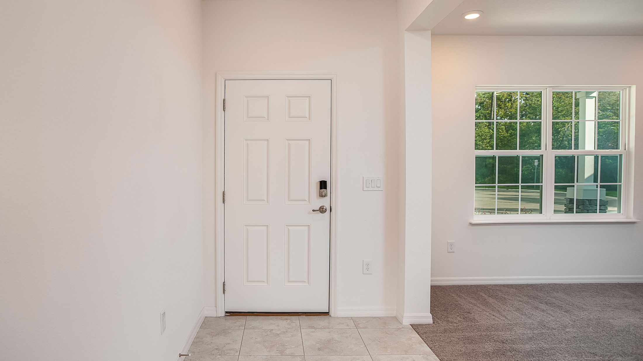 Modern interior entryway with a white door, tiled floor, and large window with greenery outside.