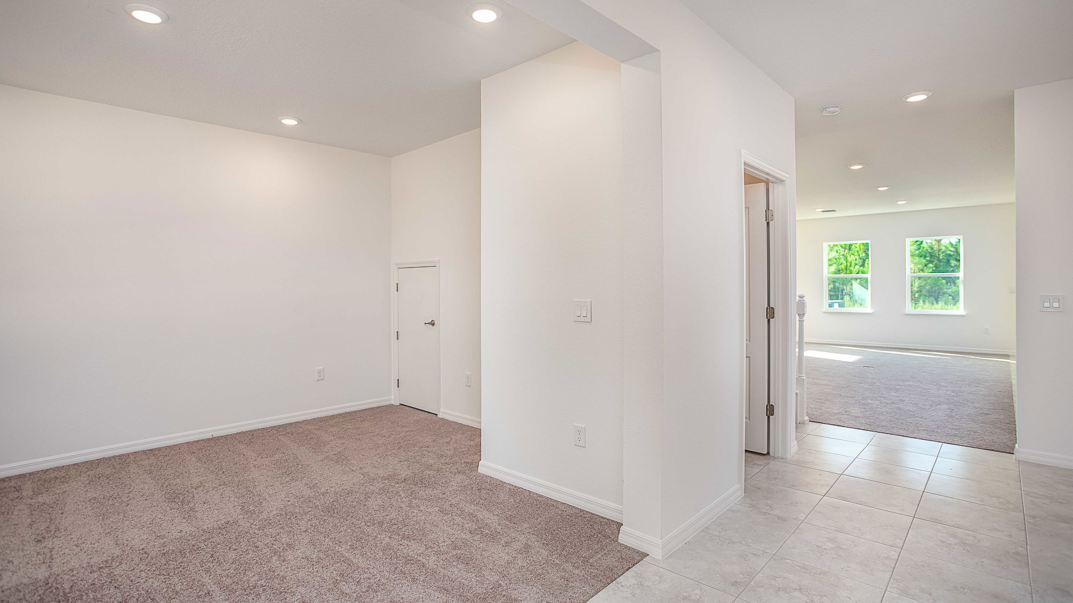 Open-concept interior of a modern home with white walls, carpeted and tiled flooring, and large windows providing natural light.