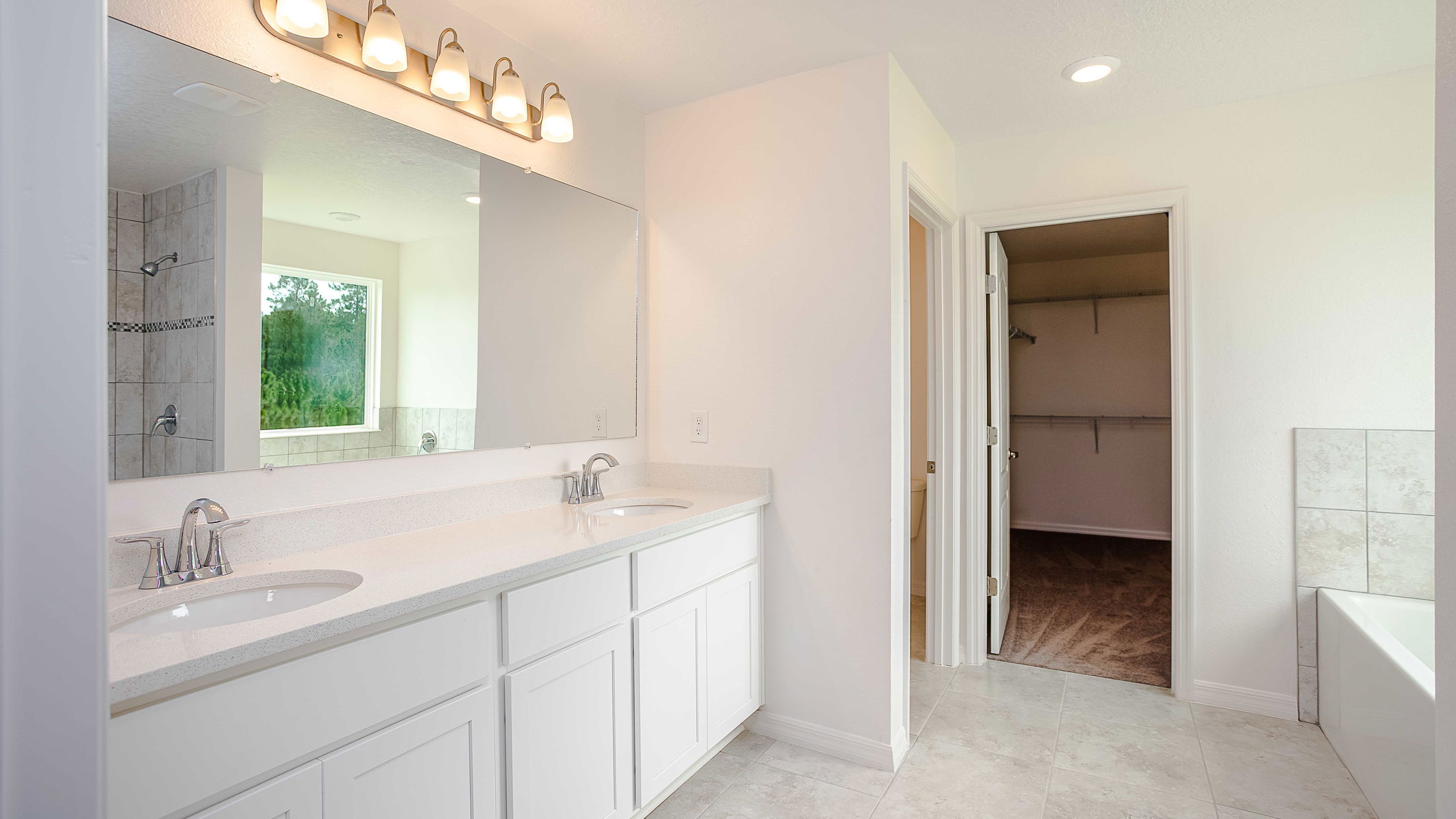 Modern bathroom interior featuring a double vanity with white cabinetry, large mirror, and illuminated vanity lights, leading to a spacious walk-in closet.