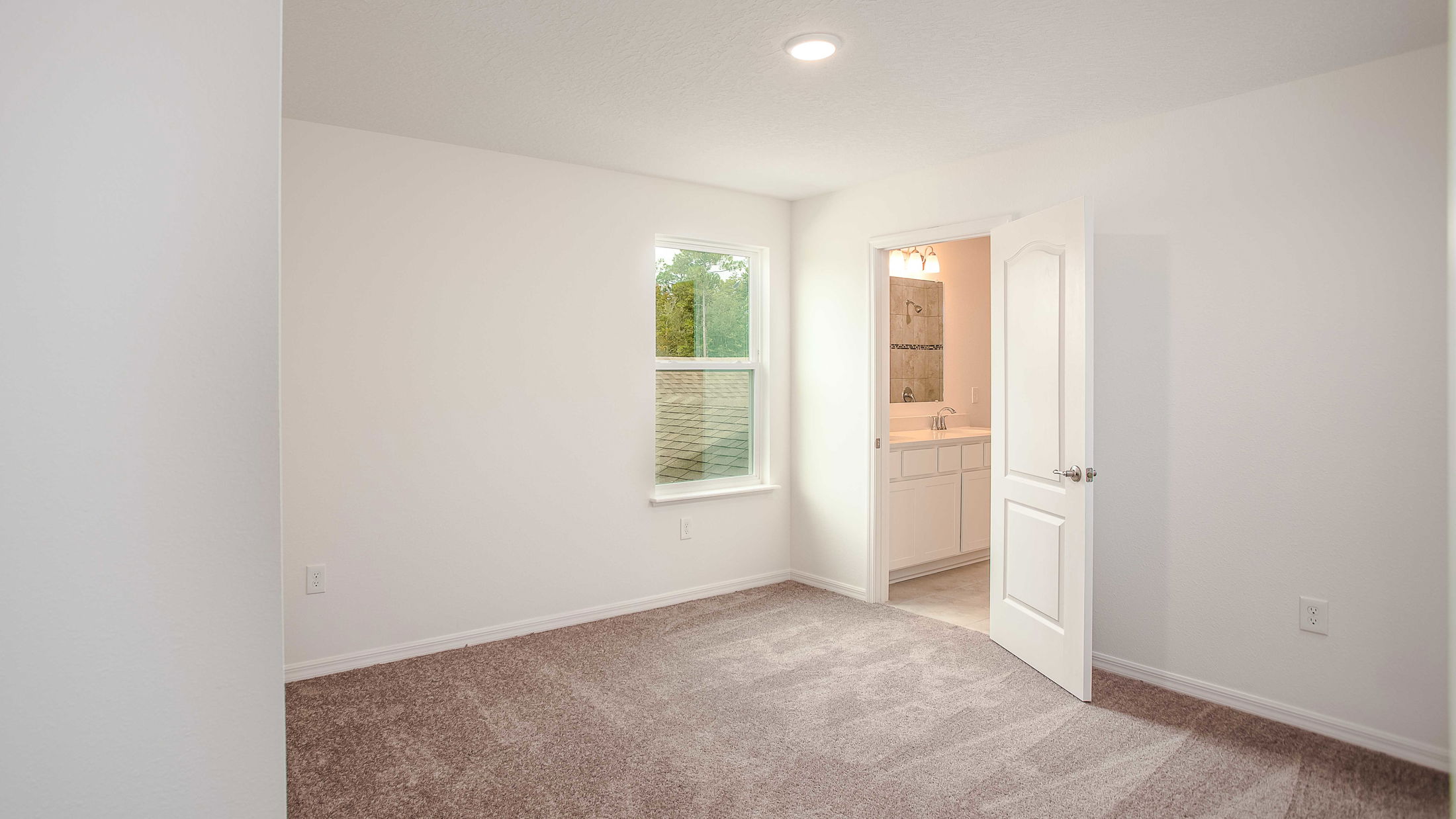 Vacant room with carpet flooring, a window, and an open door leading to a bathroom with a white vanity.