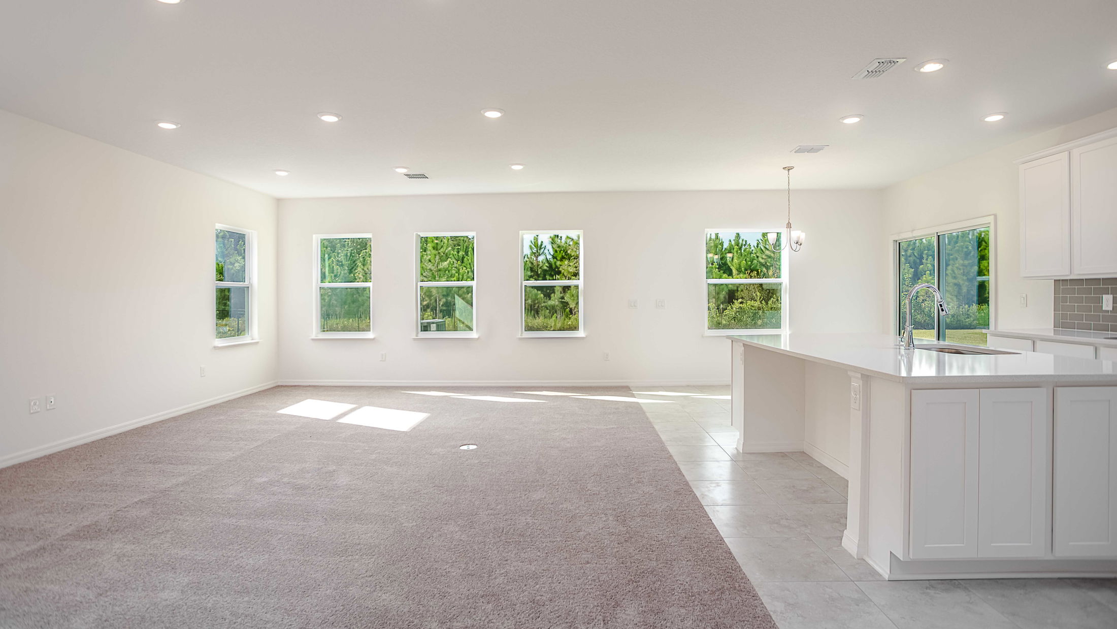 Spacious modern open-concept living room with large windows and a bright white kitchen featuring an island countertop.