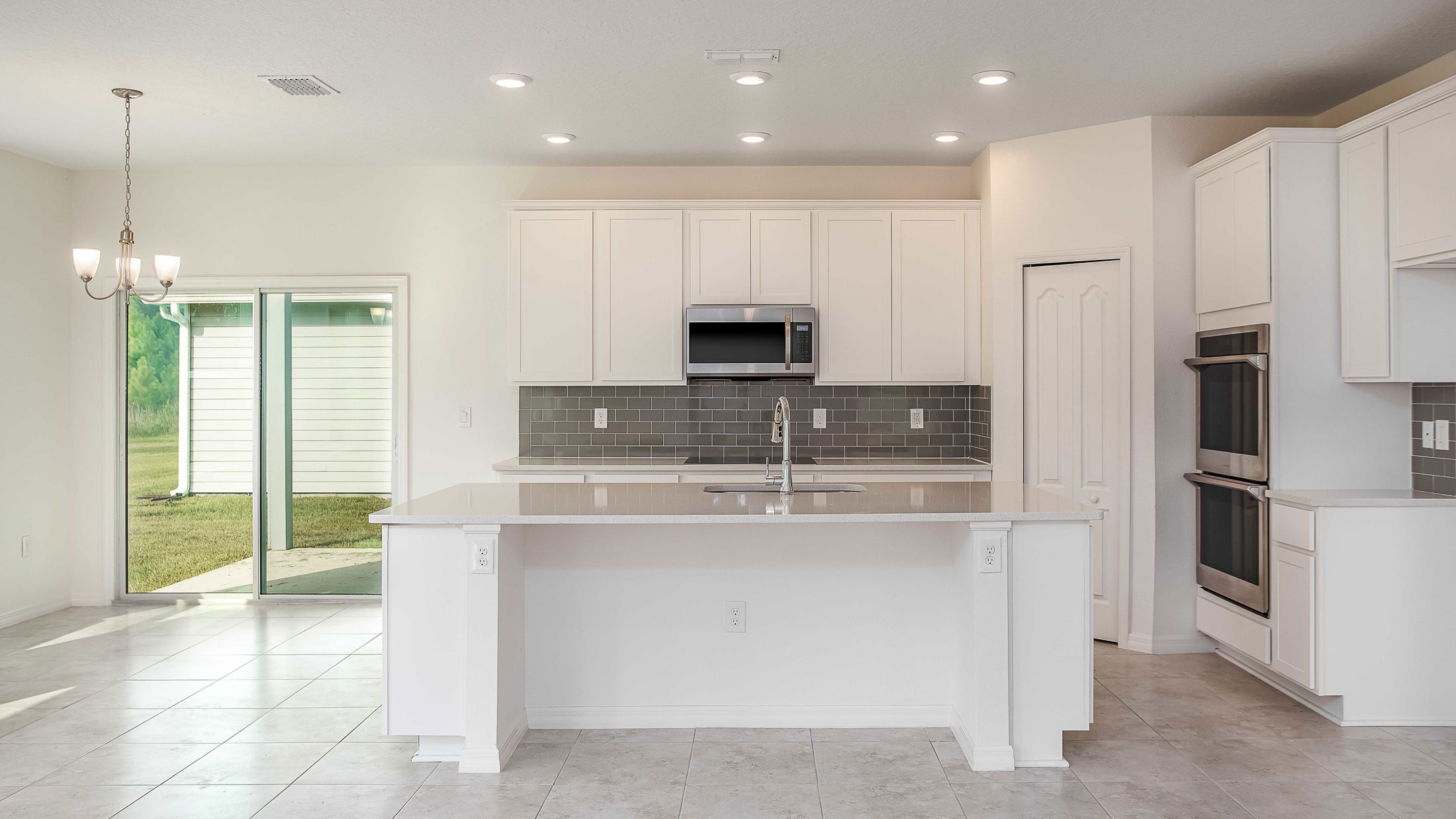 Modern kitchen interior with white cabinets, stainless steel appliances, and a central island, featuring gray subway tile backsplash and tile flooring, alongside a dining area with glass sliding doors.