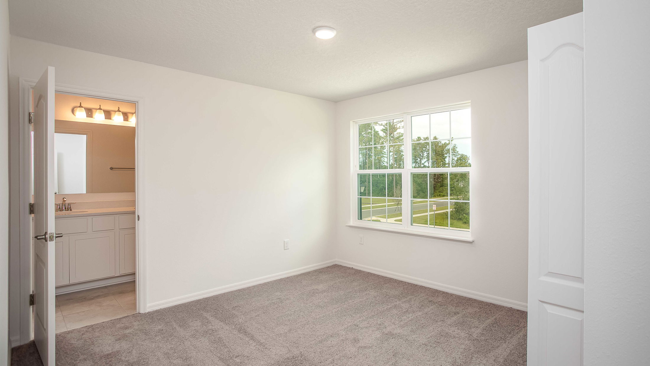Bright empty bedroom with carpet flooring, large window, and open door leading to a bathroom.
